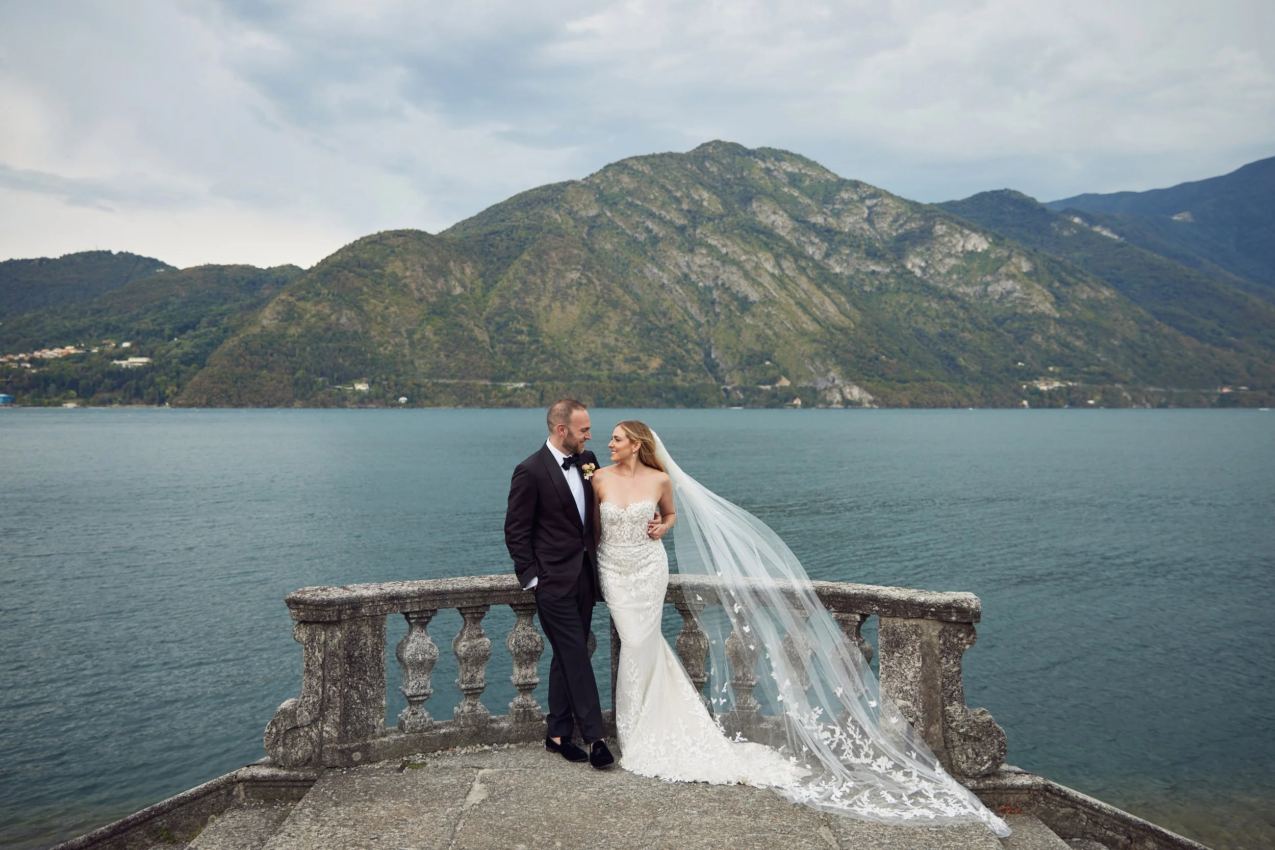 A couple in wedding attire standing by a stone railing near a body of water with mountains in the background.