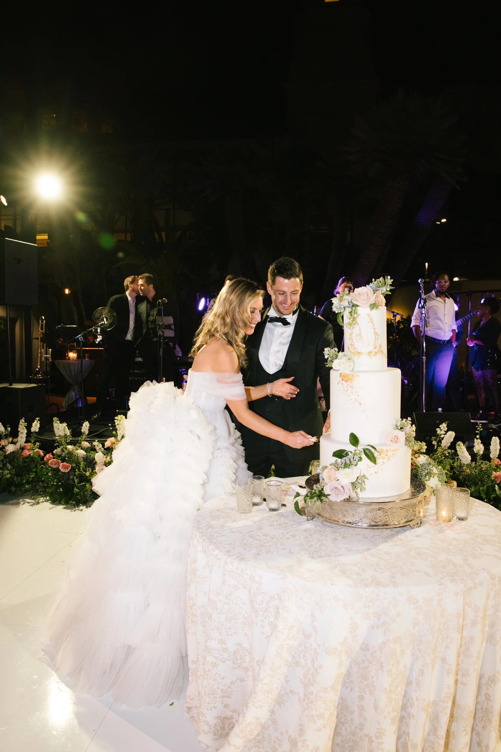A bride and groom cutting a wedding cake at their reception, decorated with flowers and surrounded by guests.