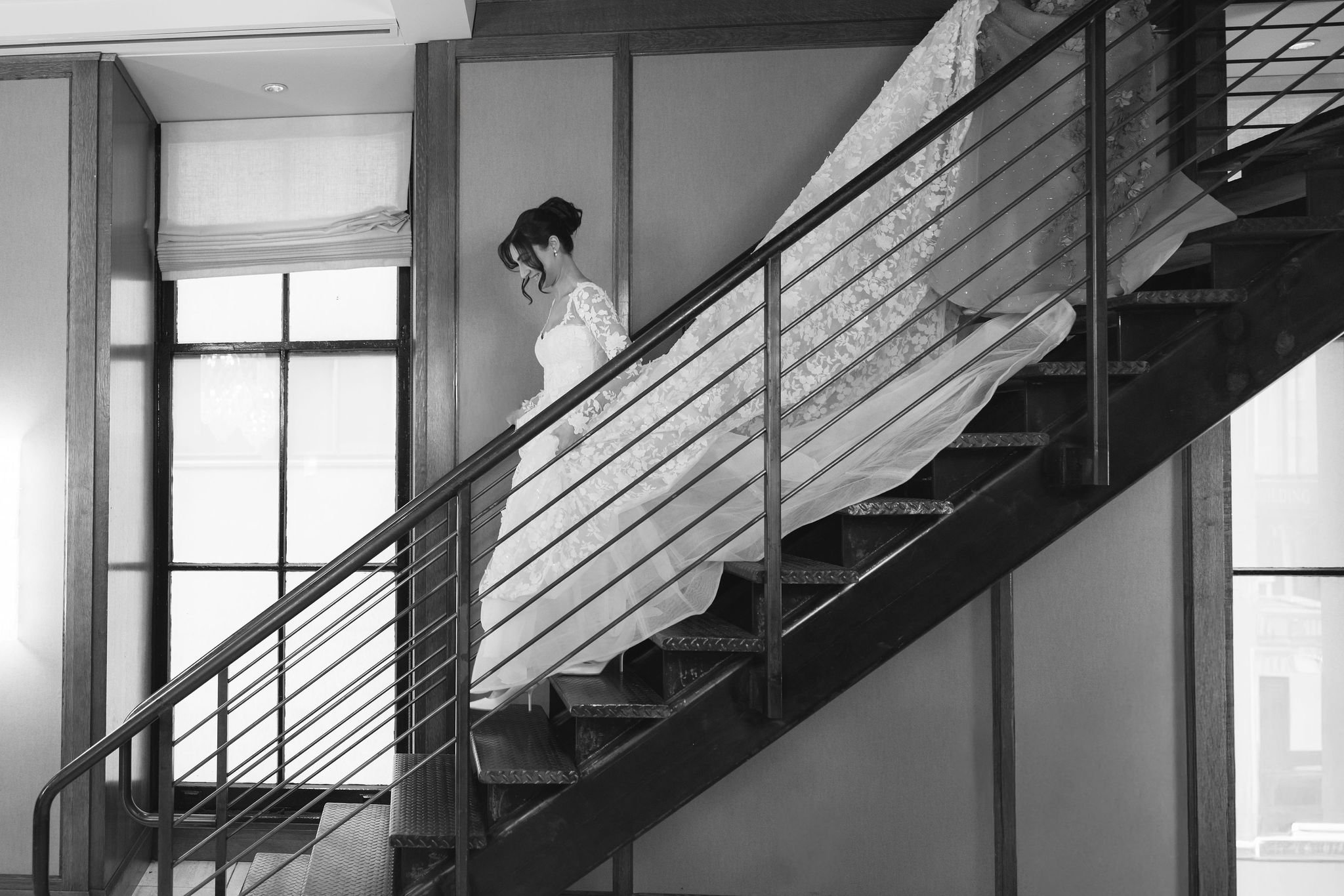 A woman in a wedding dress descending a metal staircase inside a building with large windows.