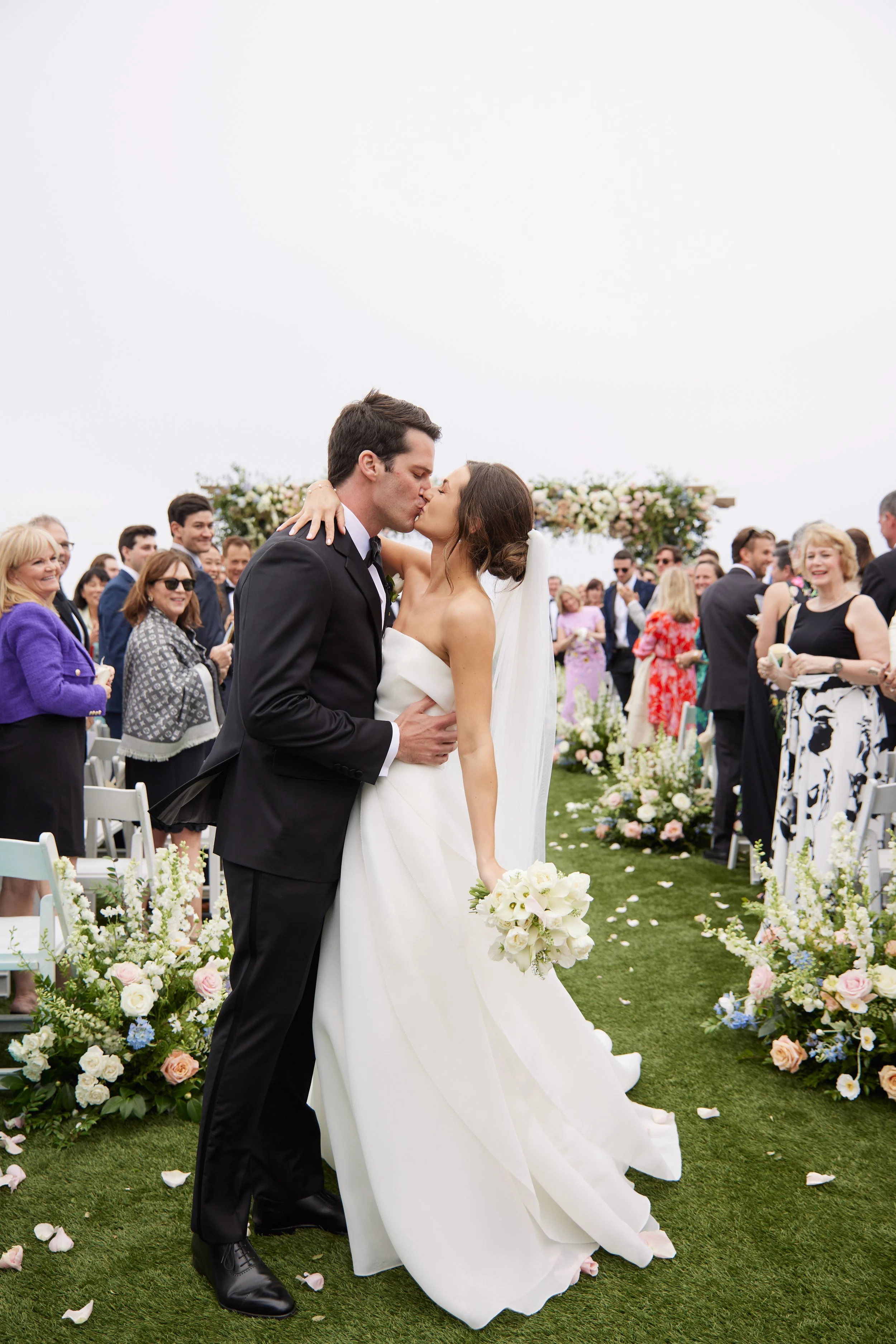 A newlywed couple kissing at their outdoor wedding ceremony, surrounded by guests and floral decorations.