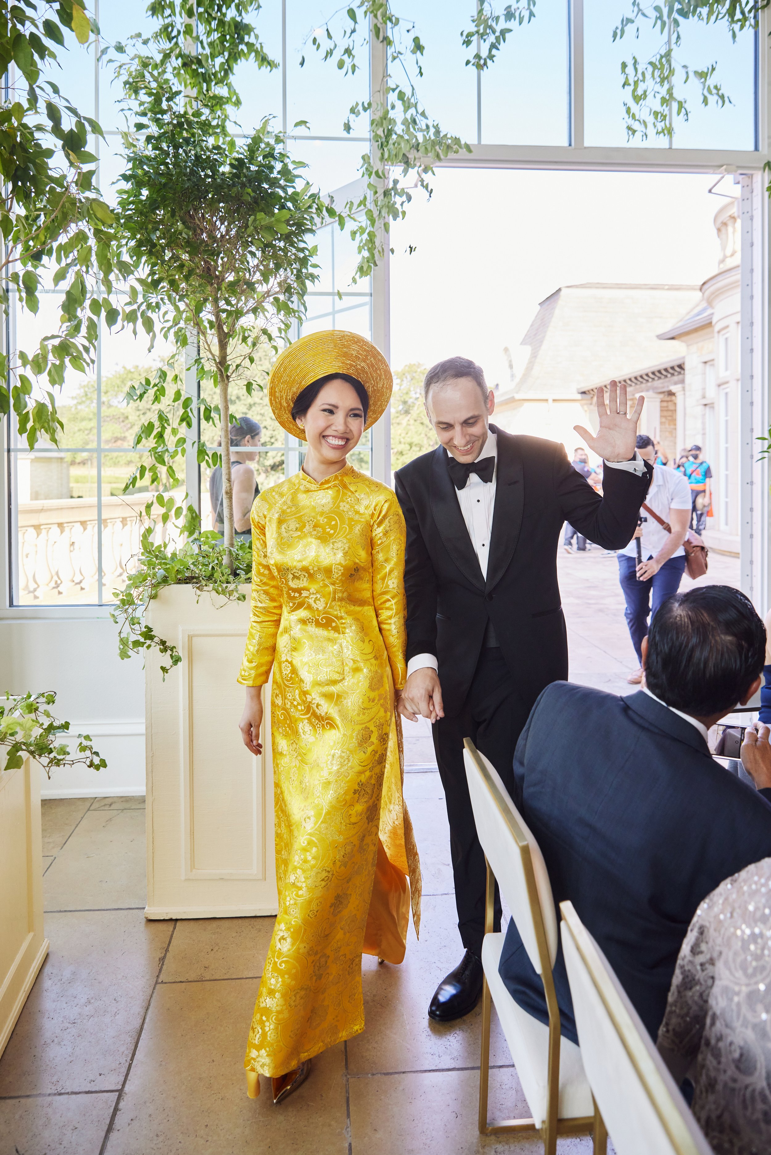 A woman in a yellow traditional dress and a wide-brimmed hat smiling as she walks through a bright indoor space, holding hands with a man in a tuxedo. There are plants and large windows in the background with people gathered outside.