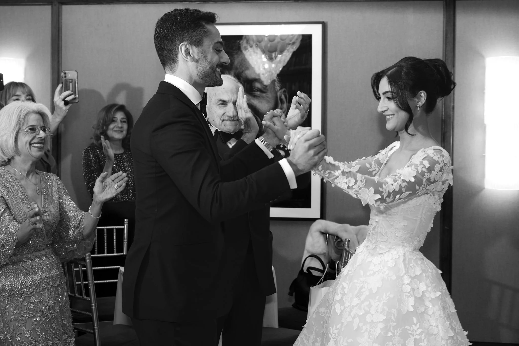 A black-and-white photo of a wedding dance with a bride and groom holding hands, smiling at each other. The groom is wearing a tuxedo and the bride is in a lace wedding gown. Several guests are visible in the background, clapping and taking pictures.