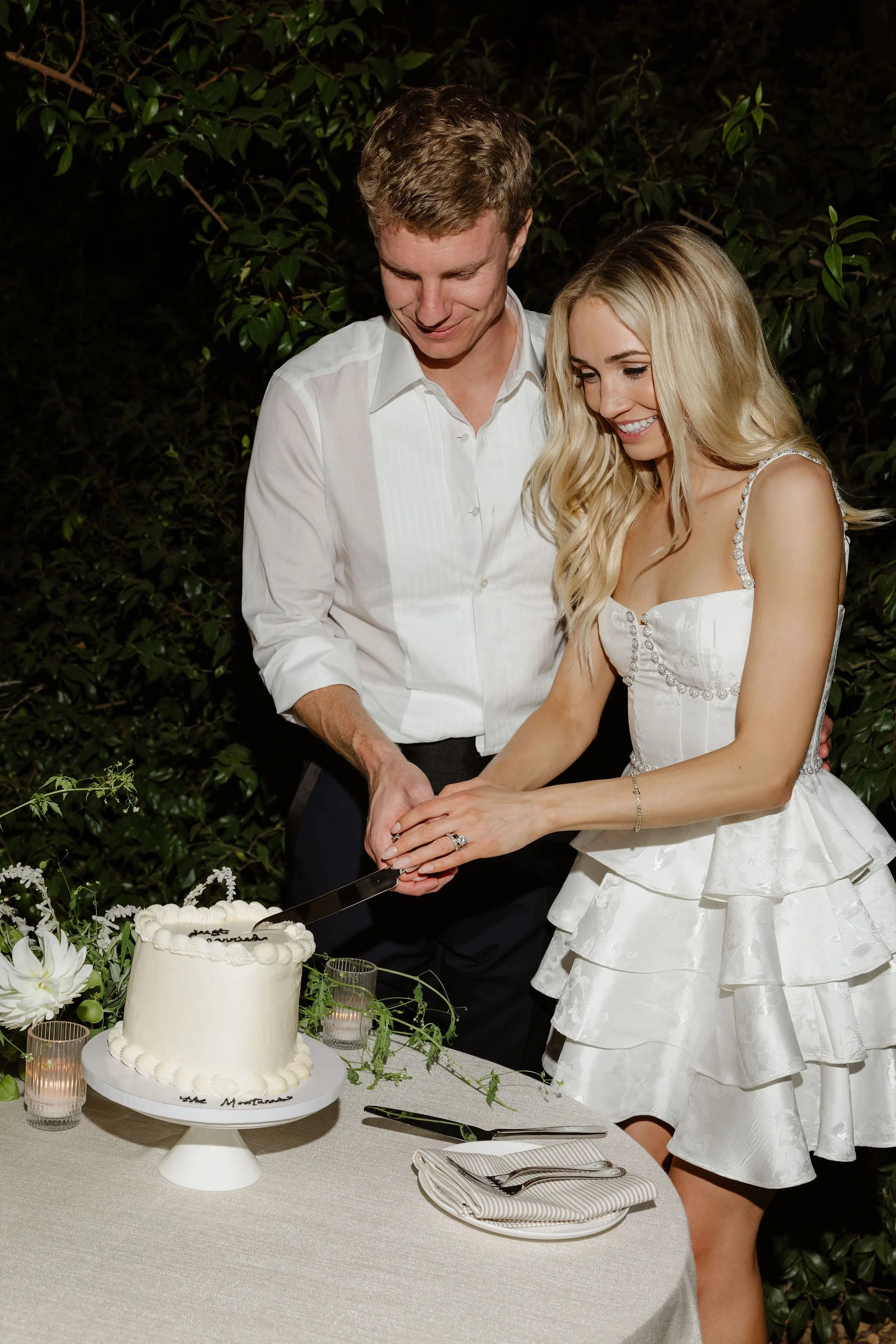 A bride and groom cutting a wedding cake together outdoors at nighttime, surrounded by flowers and candles.