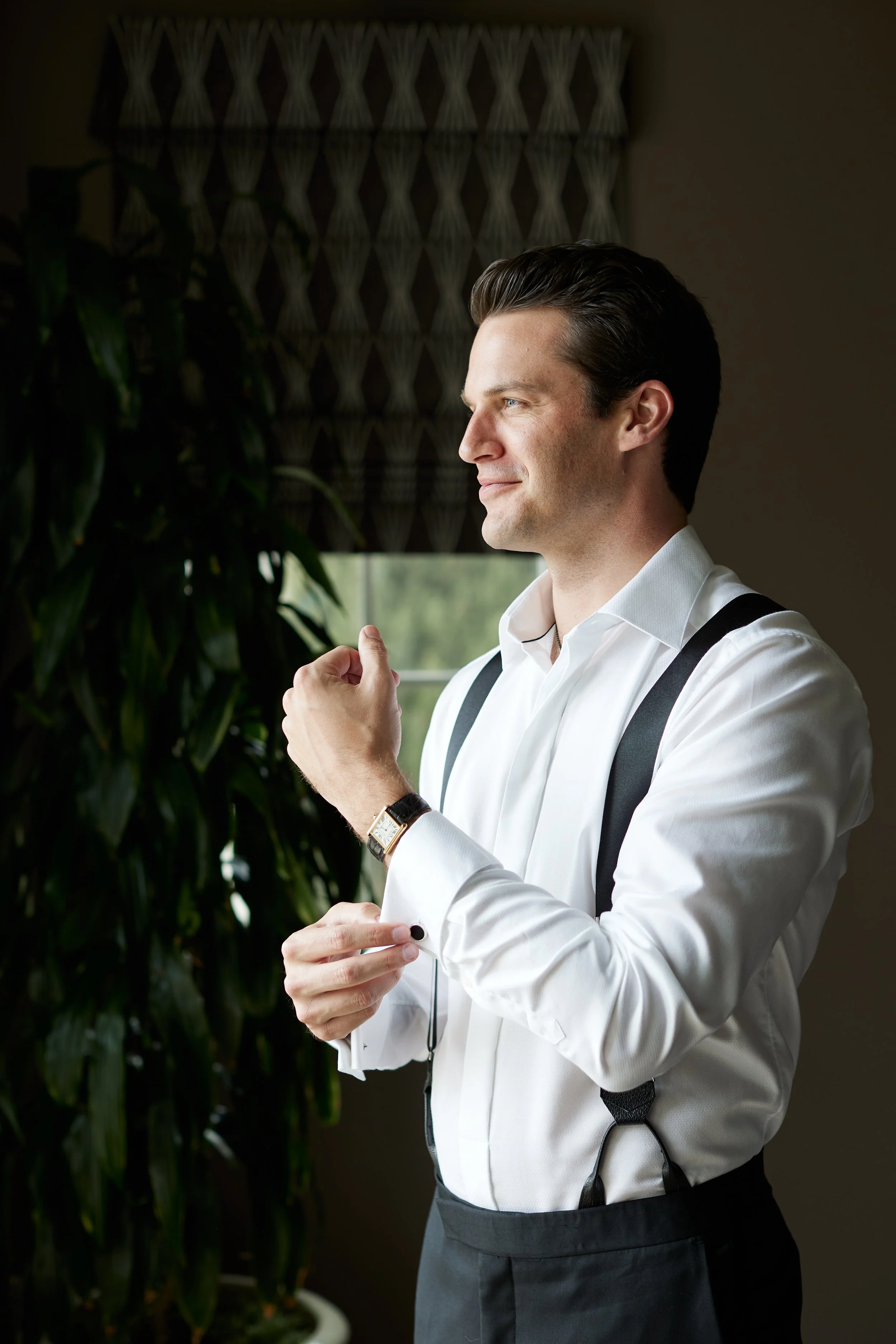A man dressed in a white shirt, gray pants, and suspenders, standing indoors near a window with dark patterned curtains and a plant, adjusting his cufflinks.