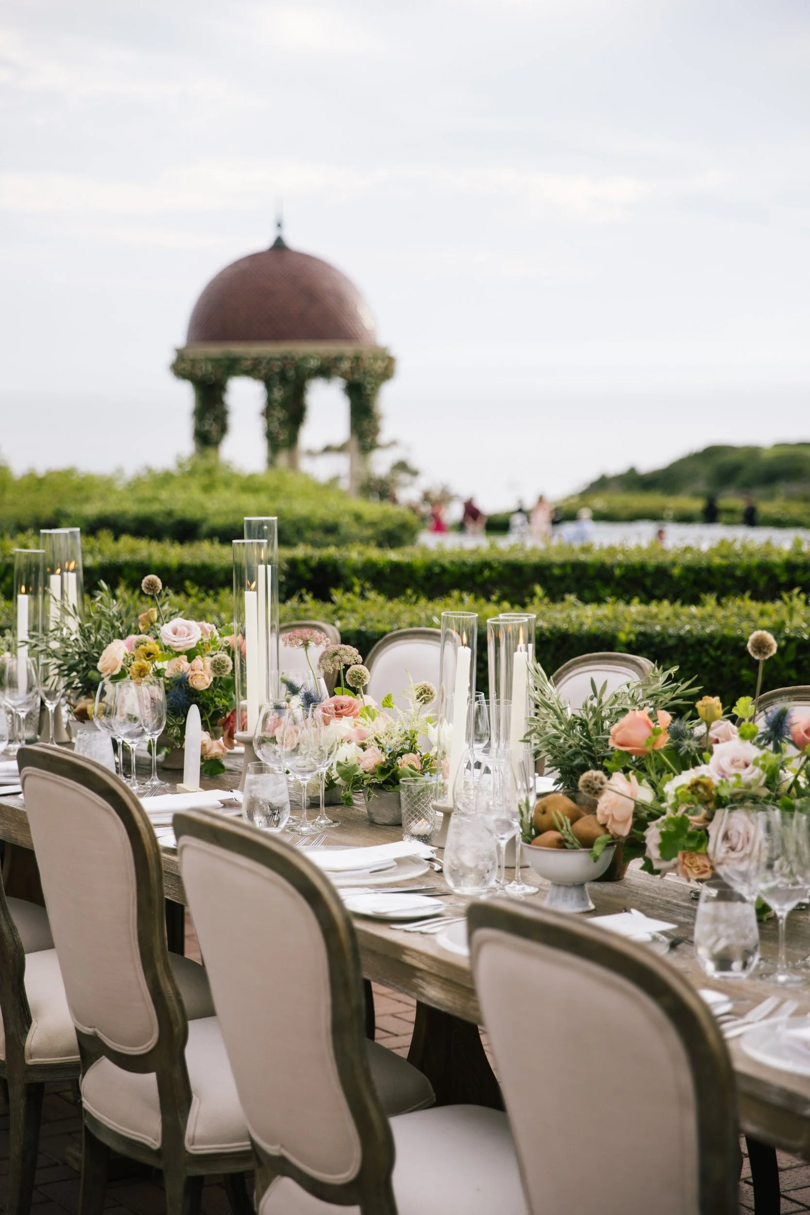 A long table set for a formal outdoor wedding reception with floral arrangements, candles, and glassware, overlooking a lush green landscape with a domed building and people in the background.