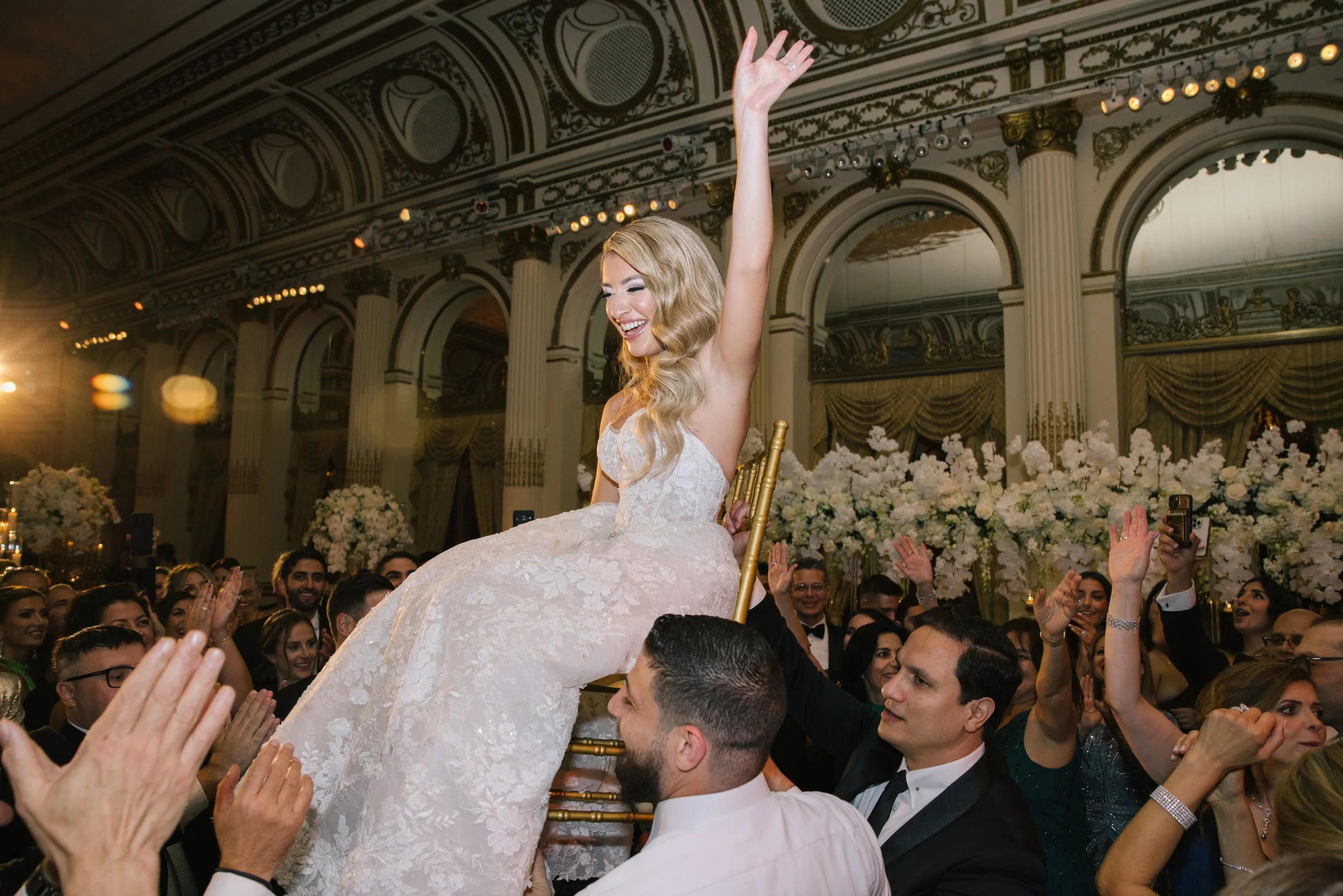 A bride with long, wavy blonde hair in a white lace wedding dress being lifted on a chair by guests at her wedding reception. She is smiling and raising one arm. The reception hall is ornate with high ceilings, columns, and floral decorations.