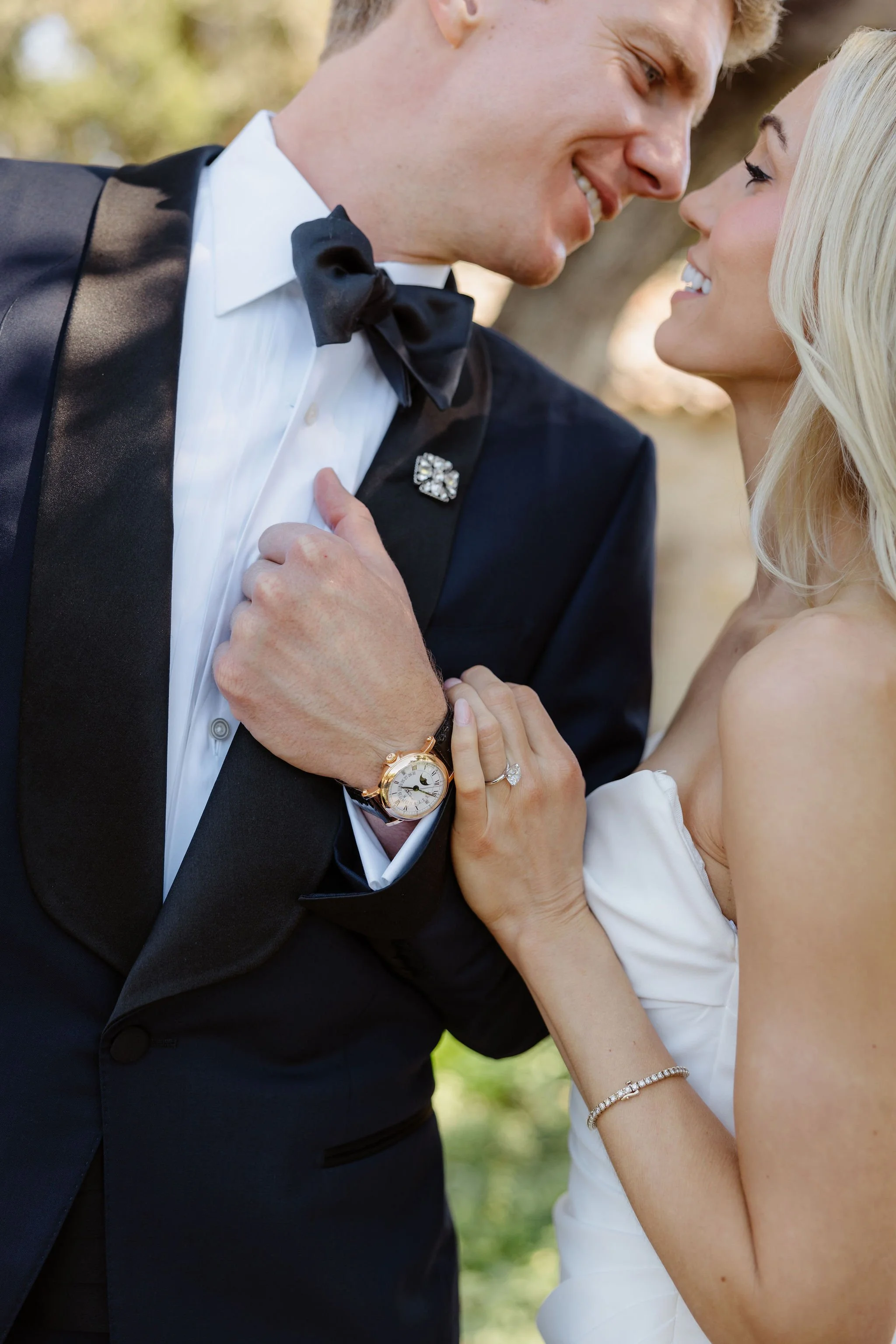 A bride and groom close together, smiling and looking at each other outdoors. The groom wears a tuxedo, and the bride is in a strapless white wedding dress, with jewelry and a bracelet. Their faces are close, and they hold each other's hands.