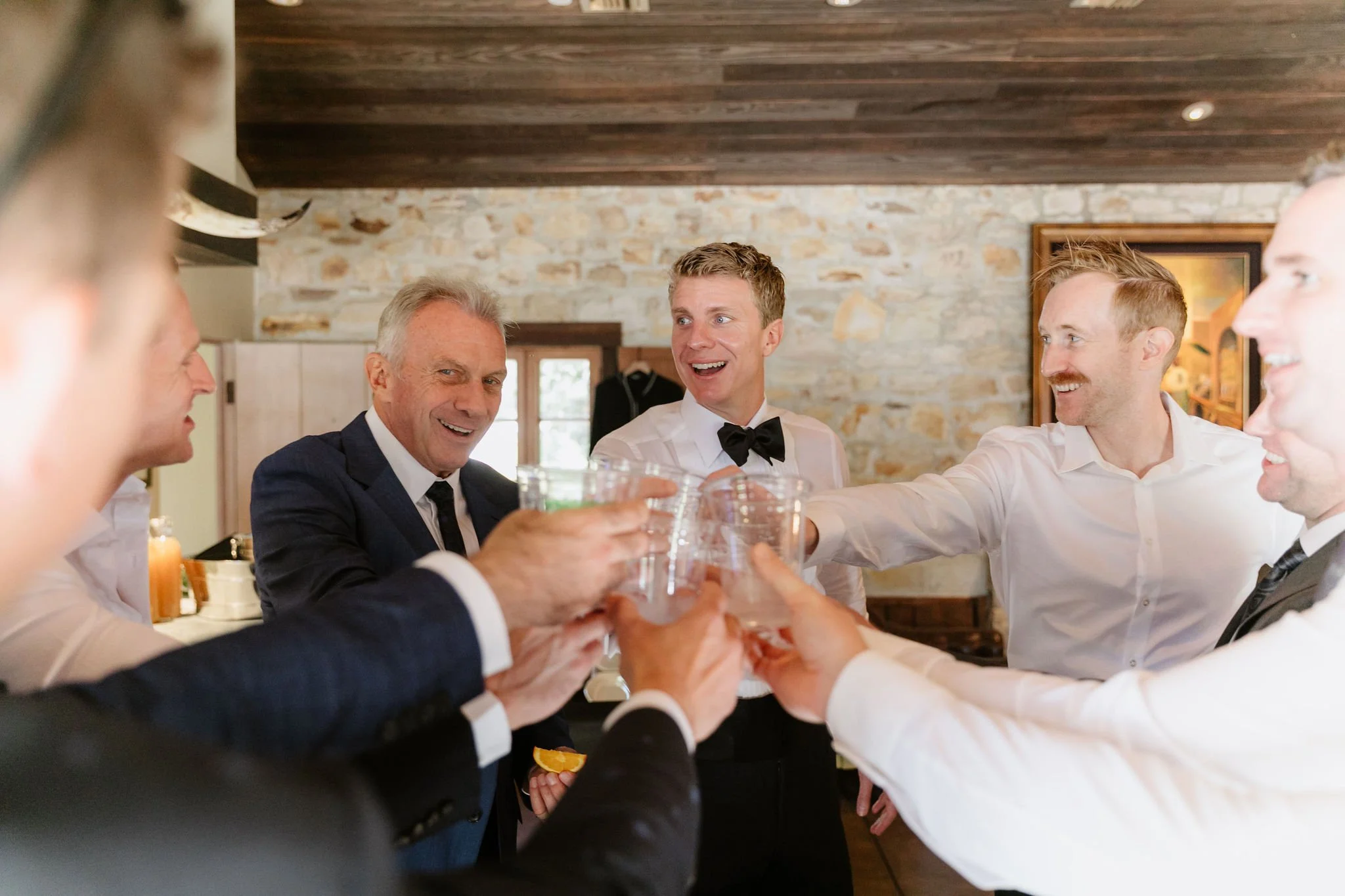 Group of men in suits and formal attire raising glasses in a toast at a celebration or gathering in a cozy, rustic-style room with a stone wall and wooden ceiling.