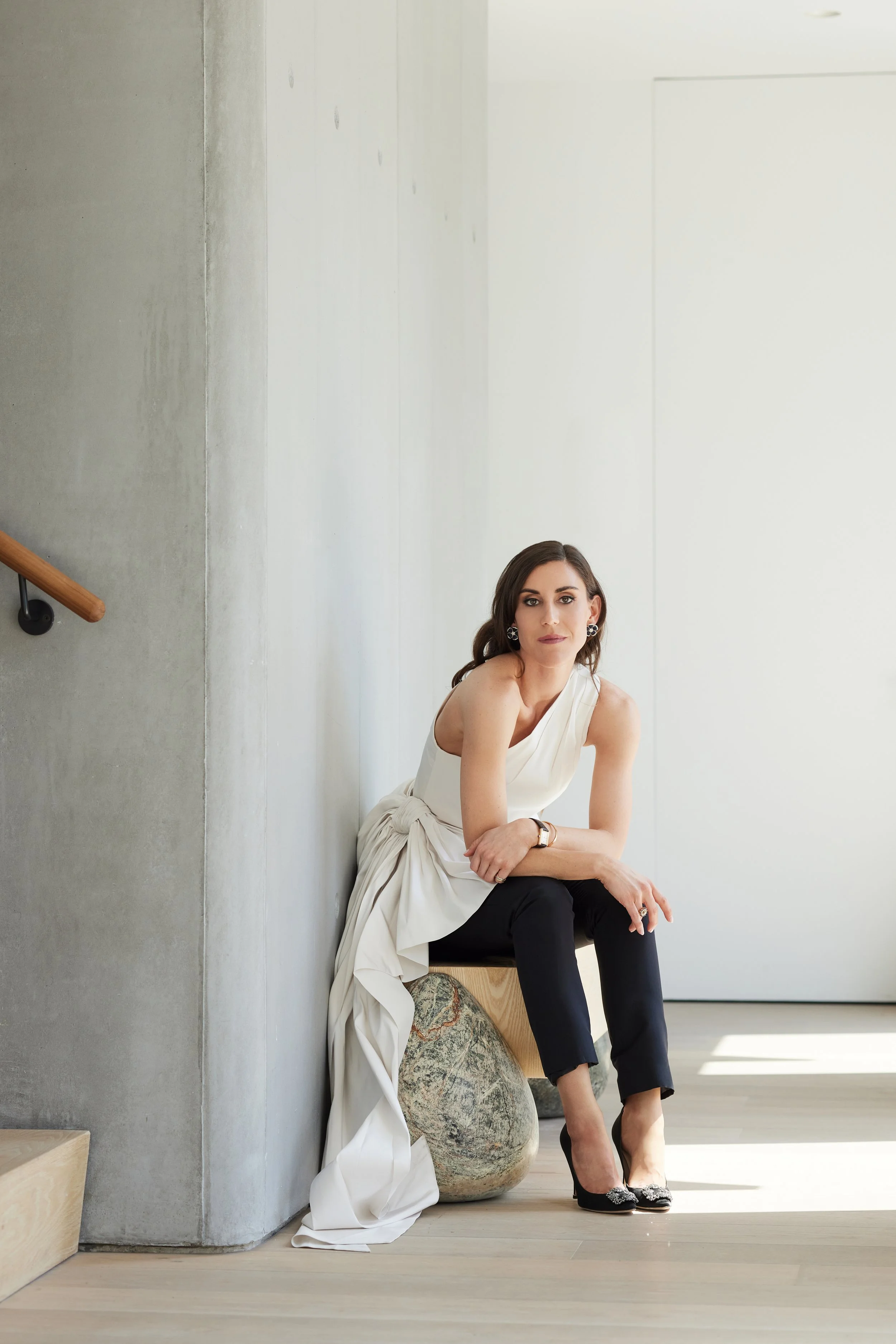 A woman in a white top and black pants sits on a bench, leaning against a concrete wall in a minimalistic, well-lit space. She has dark hair, wears earrings, and stylish shoes with embellishments.