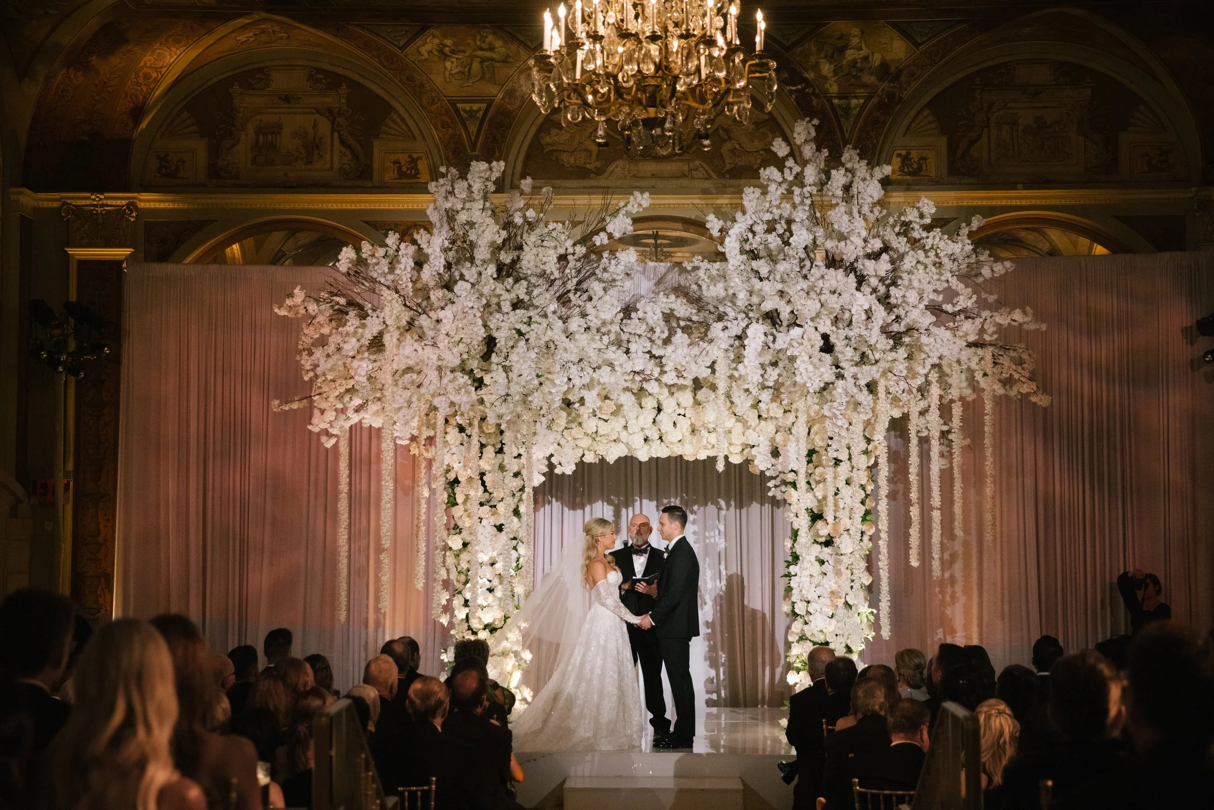 A wedding ceremony taking place in an ornate indoor venue with elaborate gold decoration, a chandelier, and floral arrangements. The bride and groom are standing at the altar, holding hands, with an officiant present.