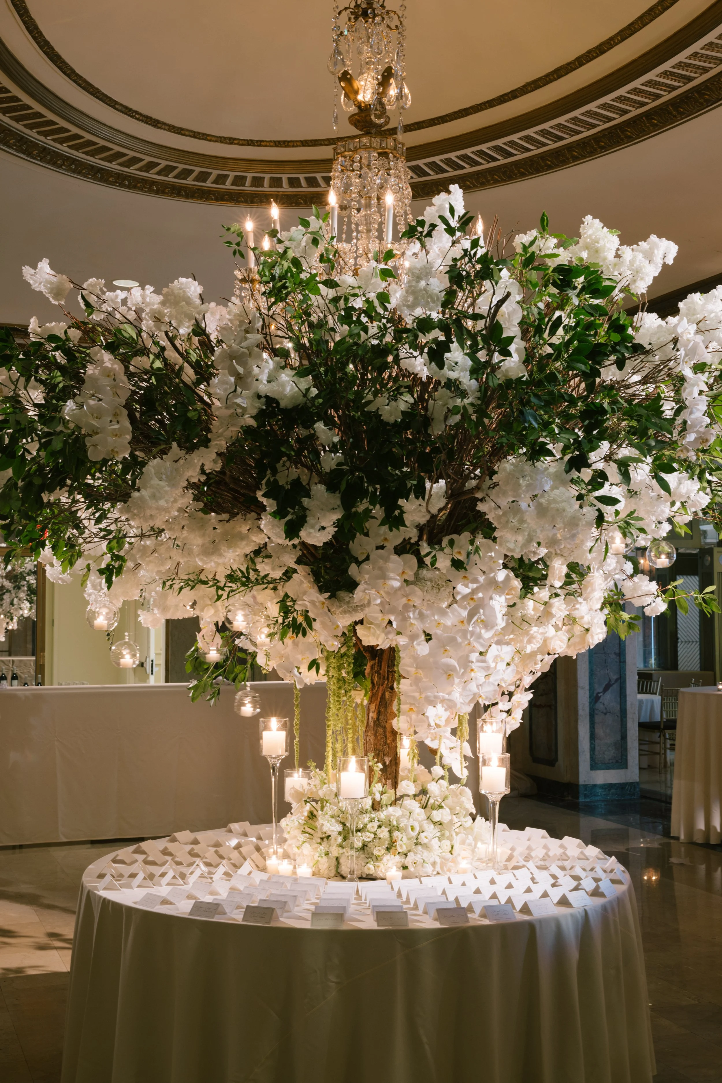 A large round table decorated with white flowers, candles, and white cards in an elegant setting, with a grand chandelier and intricate ceiling details overhead.