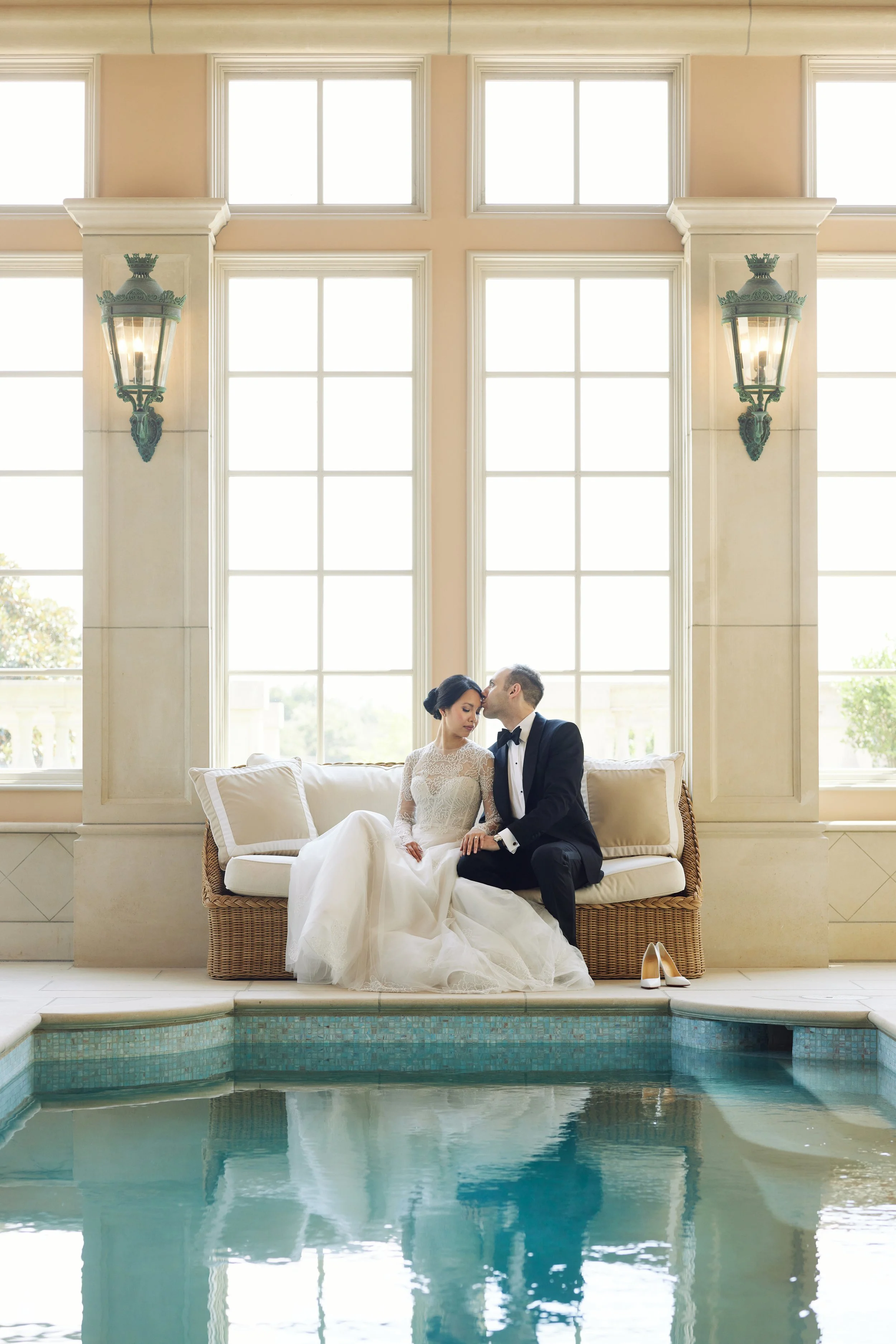 A bride and groom sitting close on a white cushioned sofa in front of large window panes, with high cream-colored walls and decorative sconces. The bride wears a white lace wedding gown, and the groom wears a black tuxedo. A pair of beige high heel shoes are on the floor beside the sofa, and a swimming pool is visible in the foreground.