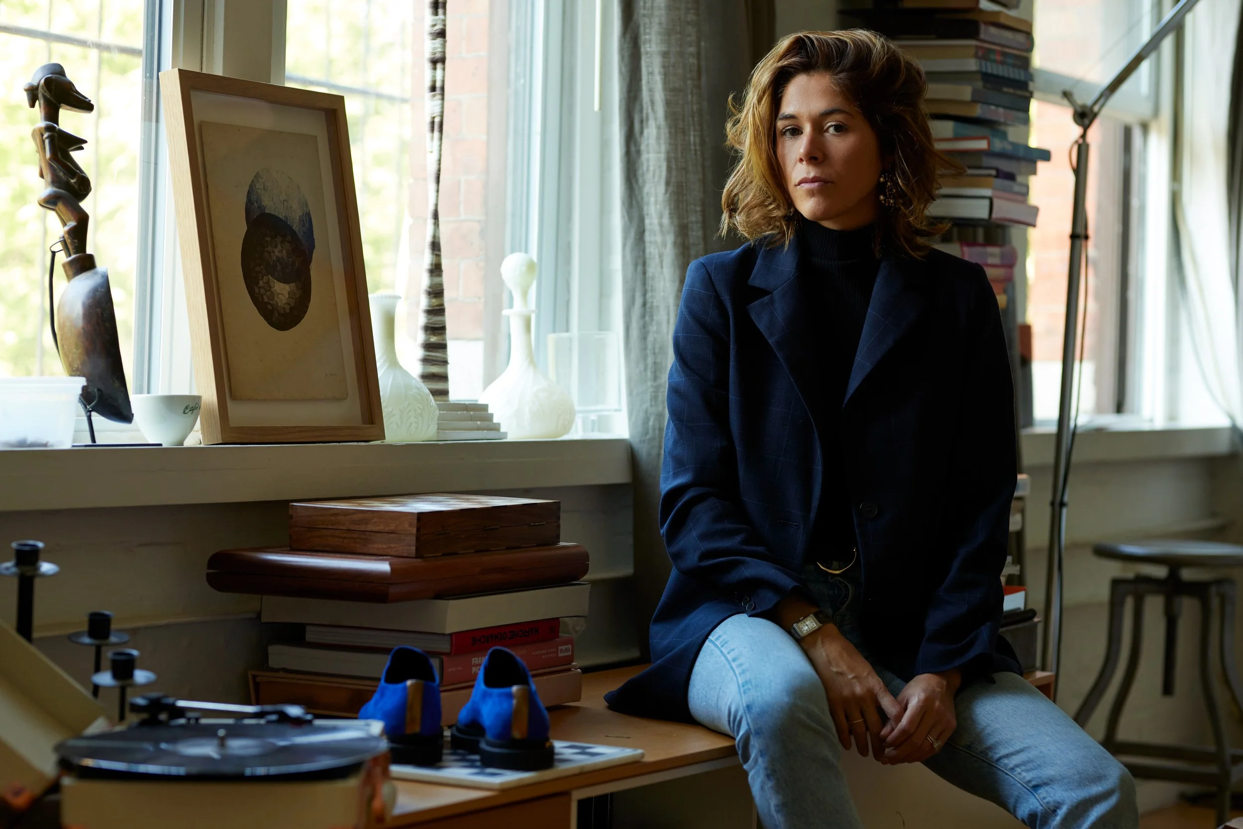 A woman with shoulder-length wavy brown hair and medium skin tone sitting in a cozy, art-filled room near a window, looking directly at the camera with a composed expression. She is wearing a dark blazer over a black top and light blue jeans. Behind 