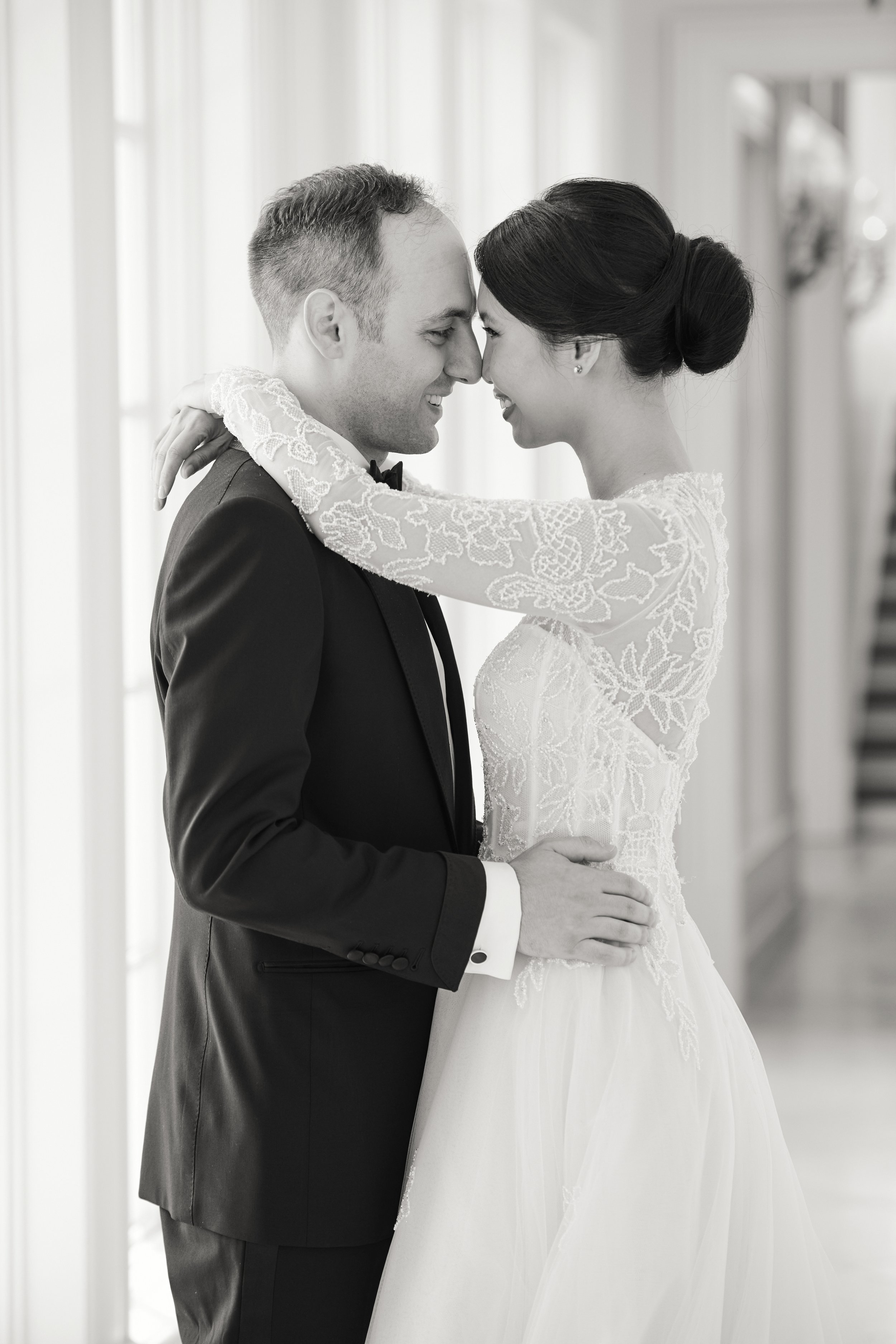 Black-and-white photo of a bride and groom touching foreheads, smiling, embracing each other in a joyful moment on their wedding day, indoors with curtains in the background.