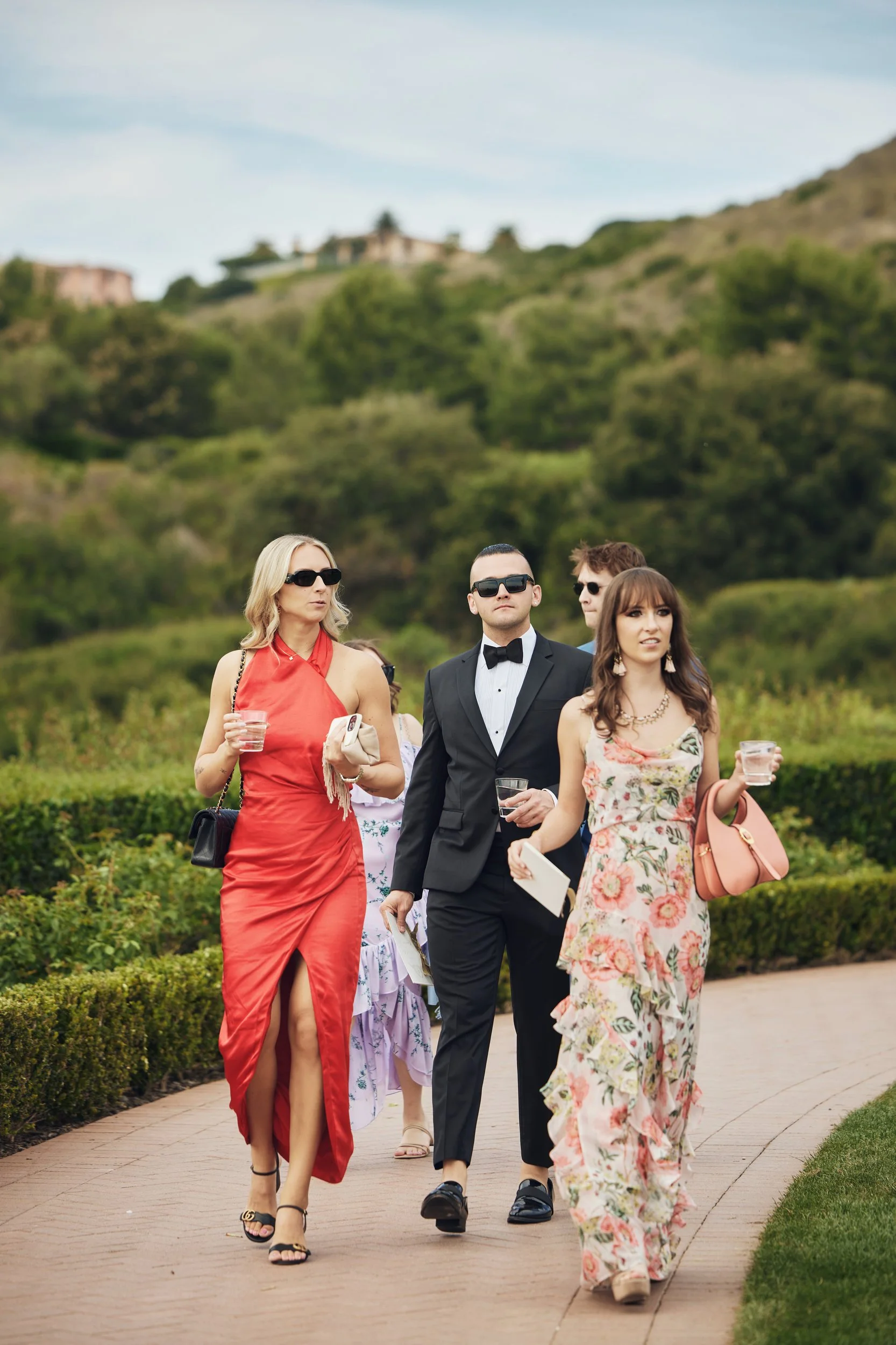 A group of well-dressed people walking outdoors on a sunny day, with a hilly landscape and houses in the background.