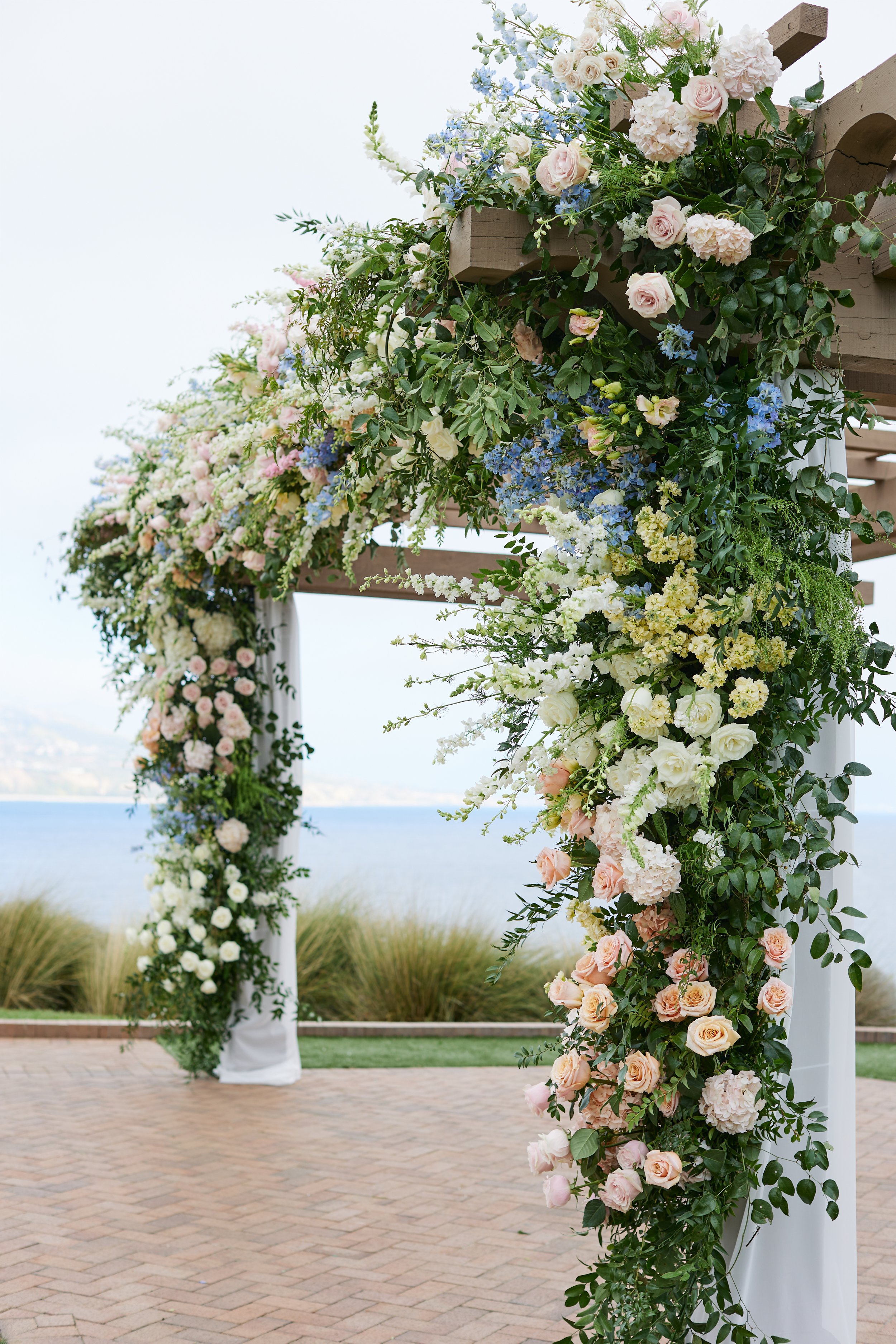 A floral wedding arch decorated with pink and white roses, hydrangeas, and greenery, set outdoors near a body of water with a brick pathway and grass in the background.
