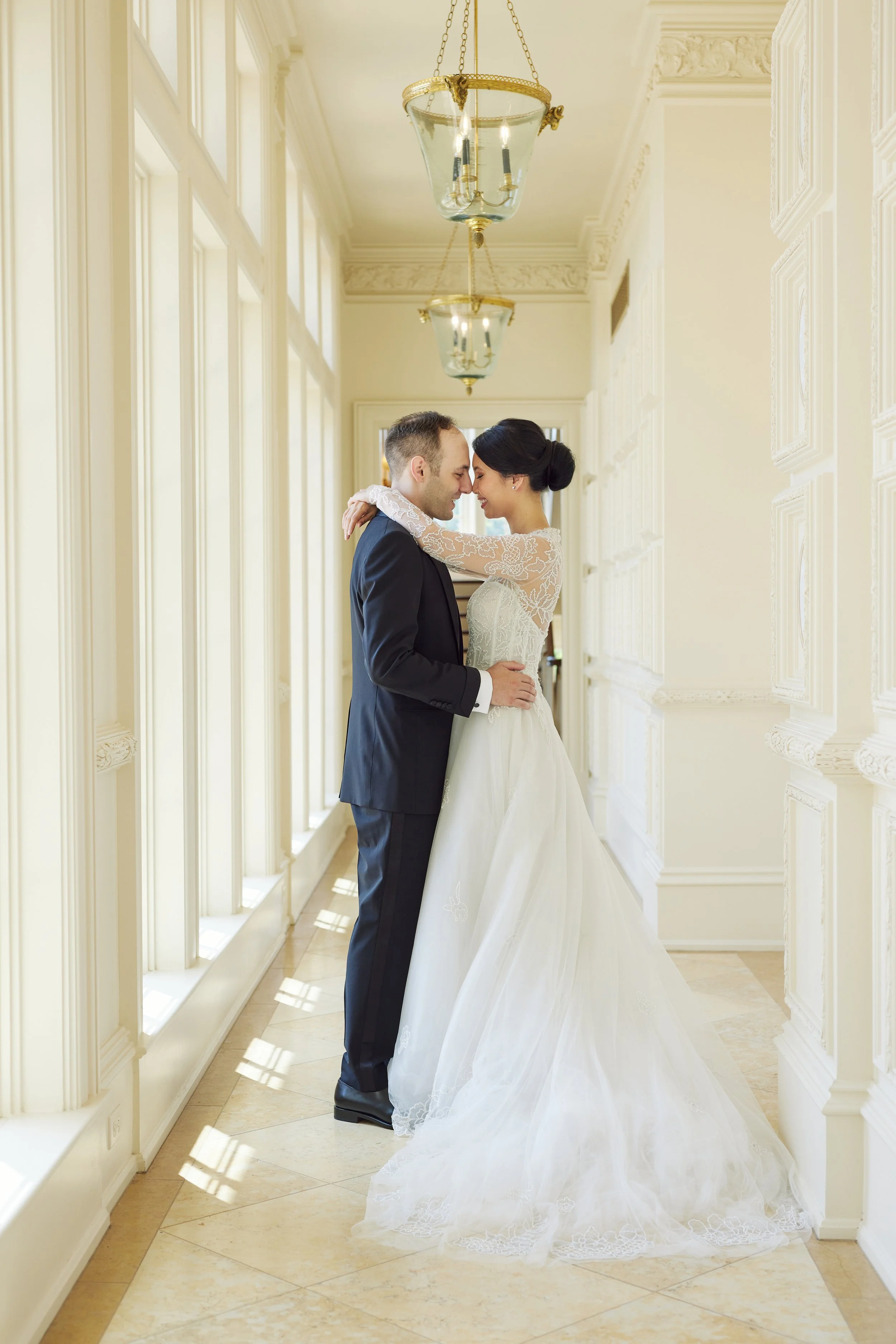 A bride and groom share a romantic moment in a bright, elegant hallway with white walls and ornate moldings. The bride wears a lace wedding gown with a long train and her dark hair in an updo. The groom is dressed in a black tuxedo with a white shirt. They are holding each other tenderly with foreheads touching.