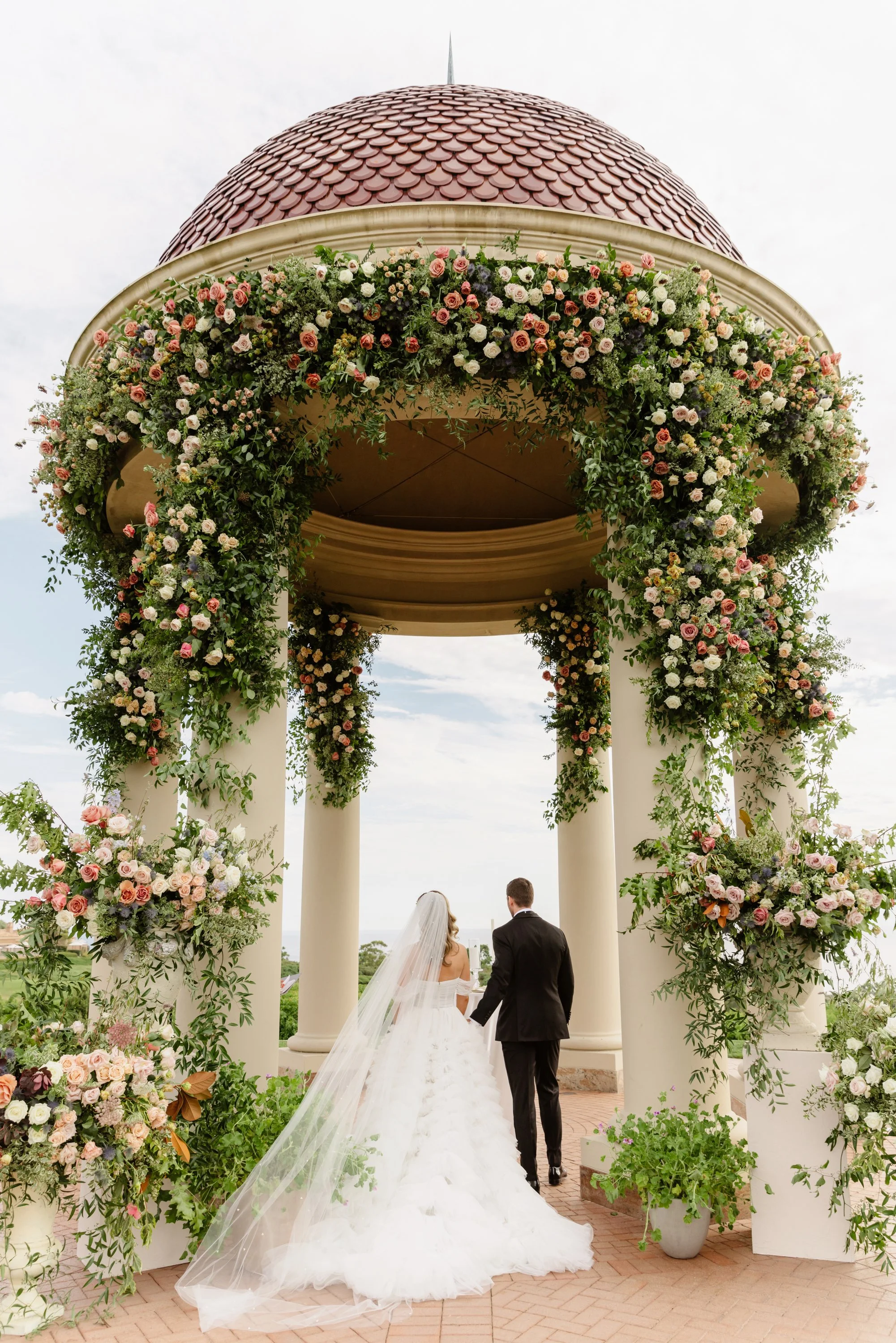 A bride and groom walking hand-in-hand through a floral archway during an outdoor wedding ceremony, with white columns and lush greenery, under a cloudy sky.