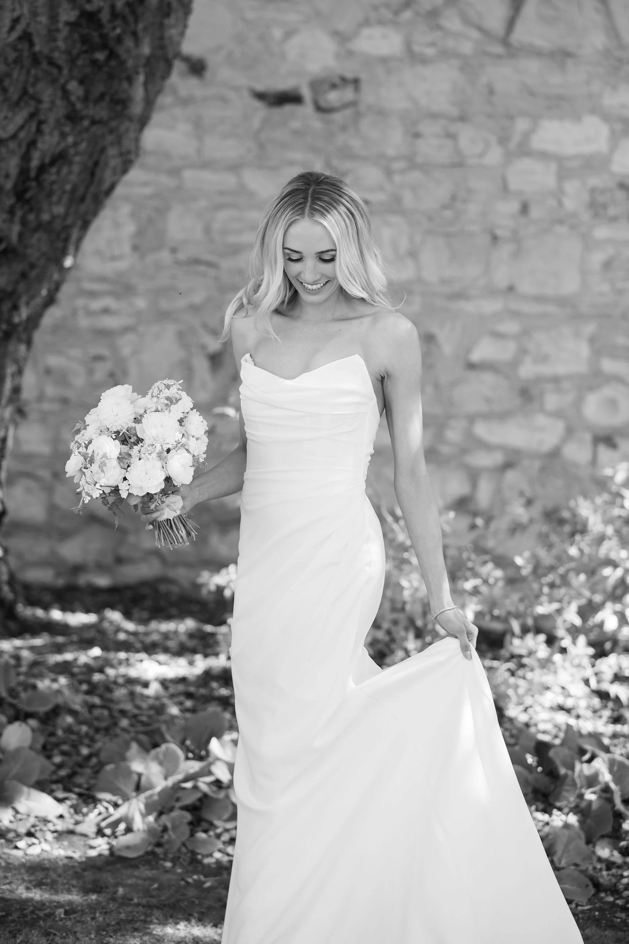 A woman in a wedding dress holding a bouquet of flowers, smiling and looking down, outdoors near a stone wall with a tree trunk nearby.