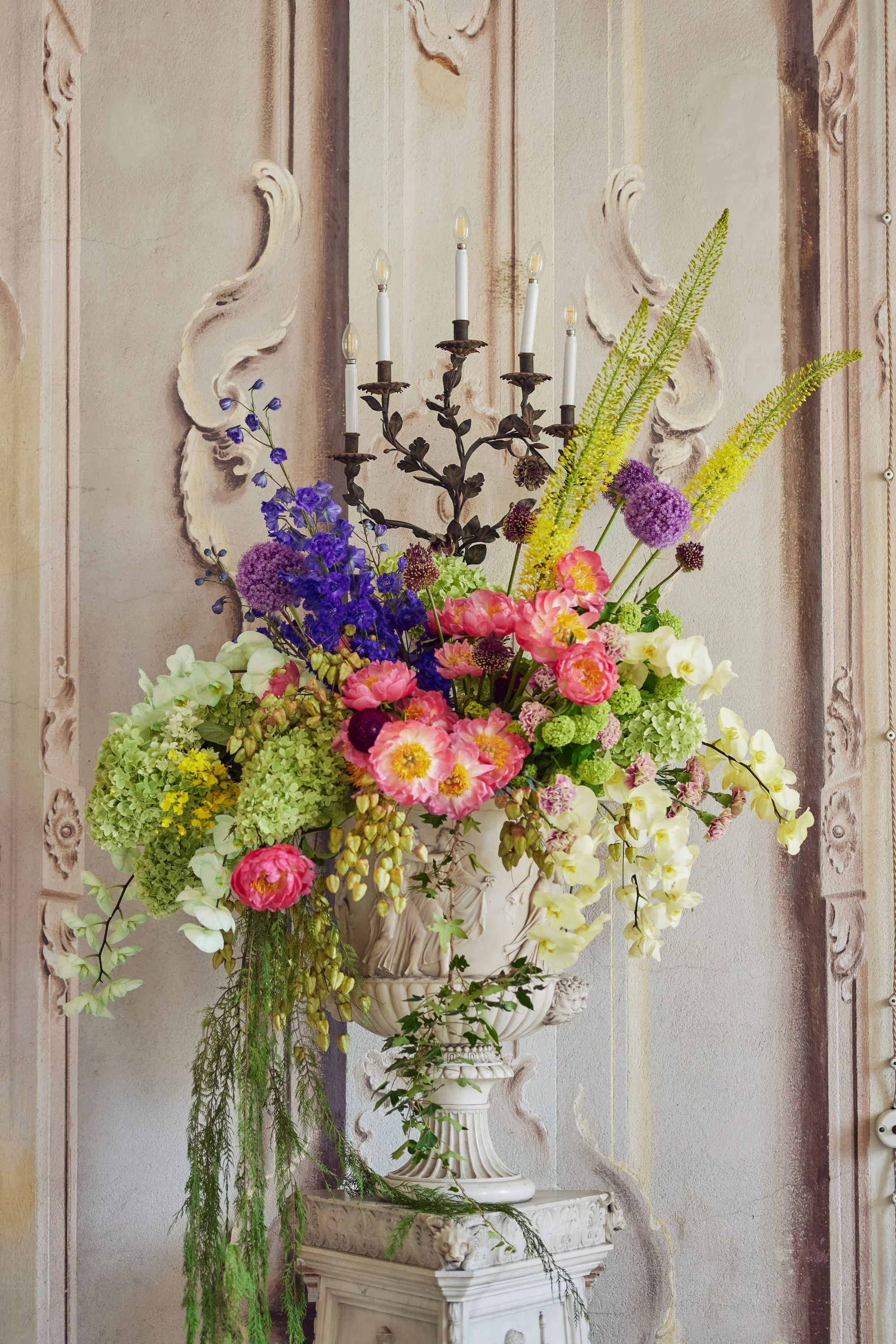 A large flower arrangement in a decorative white urn with pink, purple, green, and white flowers, including hydrangeas, poppies, and delphiniums, set on a pedestal against a wall with ornate molding and a vertical stripe design.