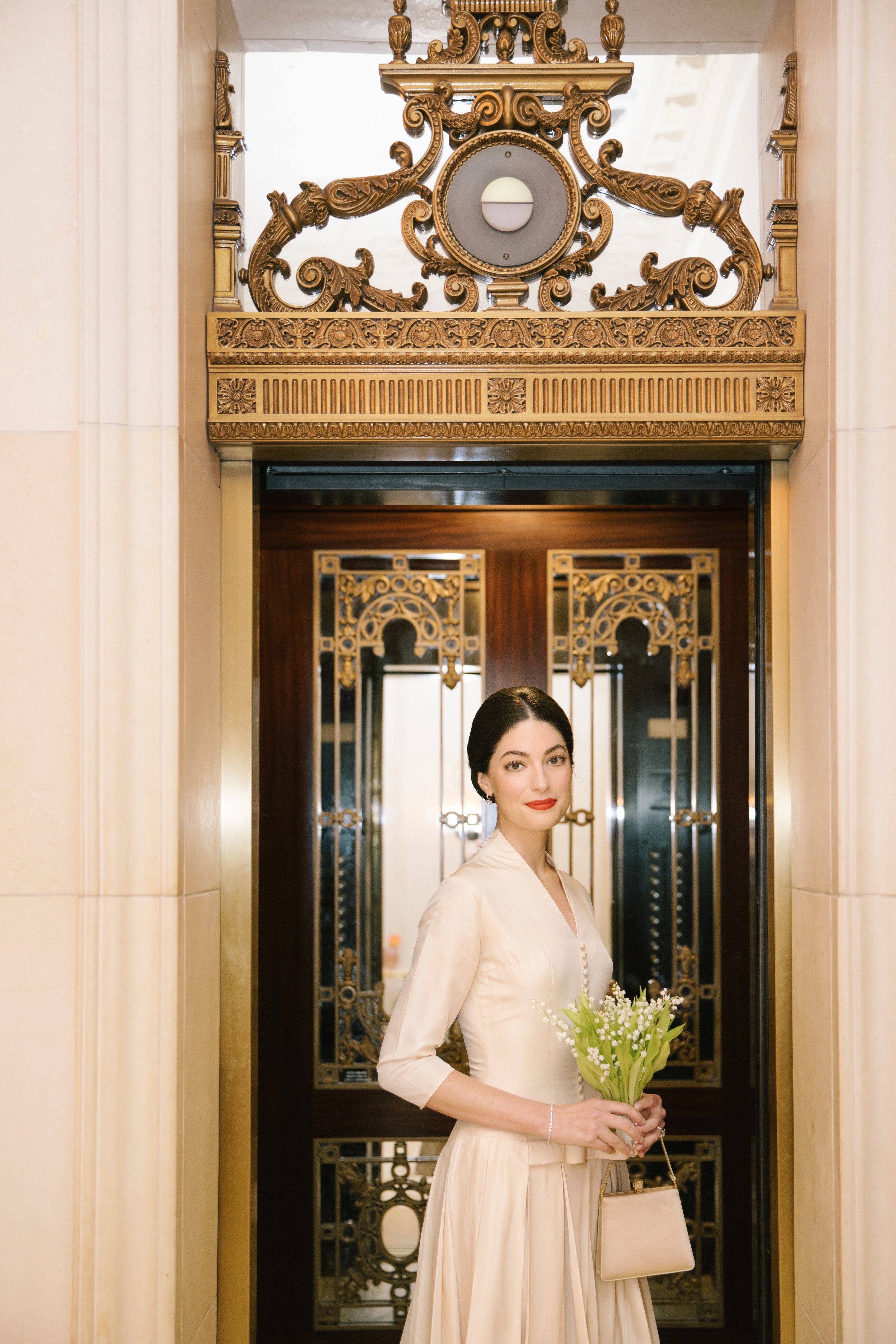 A woman in a cream-colored dress holding a bouquet of white flowers and a beige purse standing in an ornate gold and wood elevator lobby.
