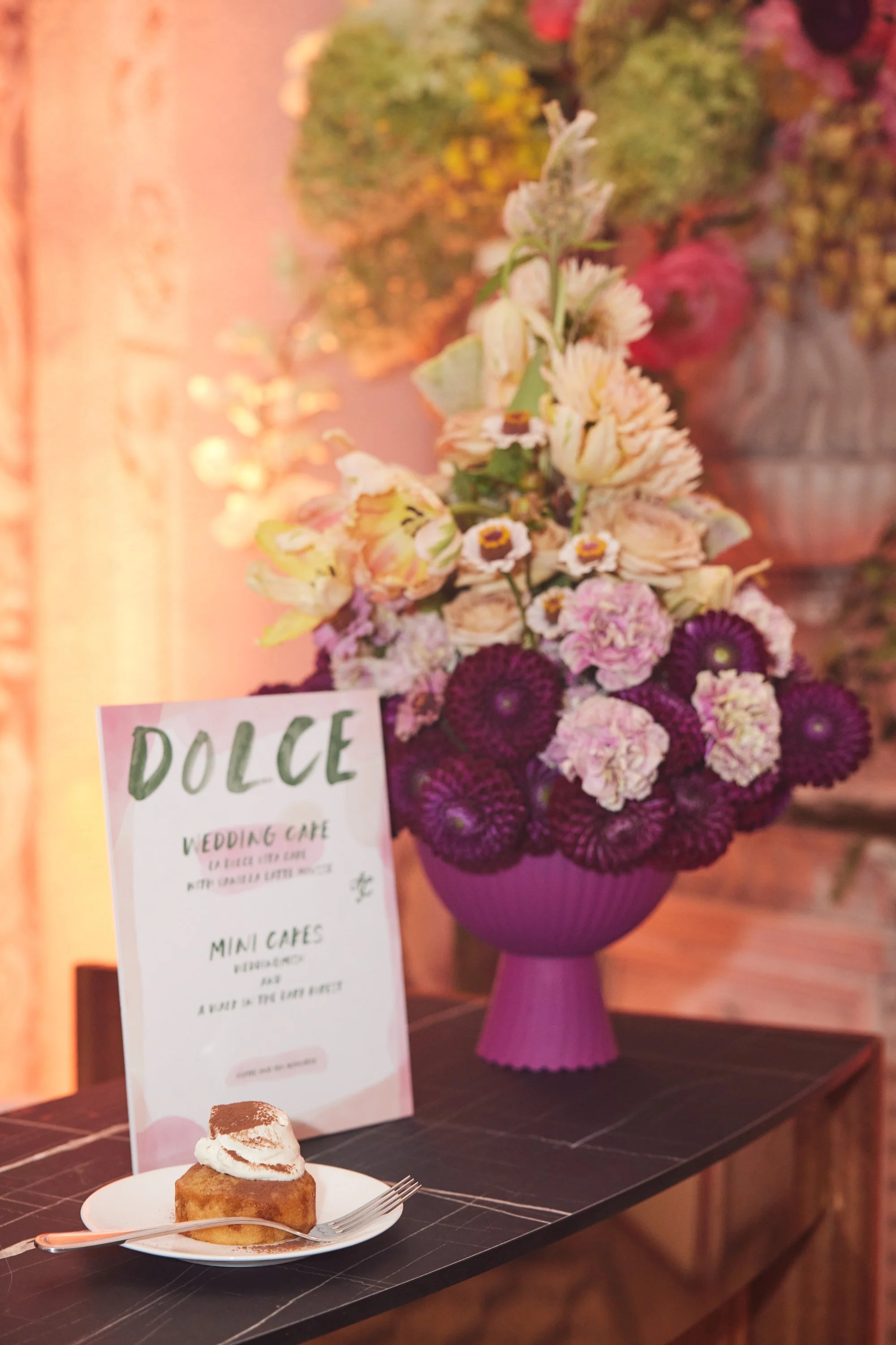 A flower arrangement in a tall purple vase with yellow, pink, and white flowers including daisies and carnations, placed on a black wooden table. A small dessert topped with whipped cream and cocoa powder is in front of the arrangement. There is a wh