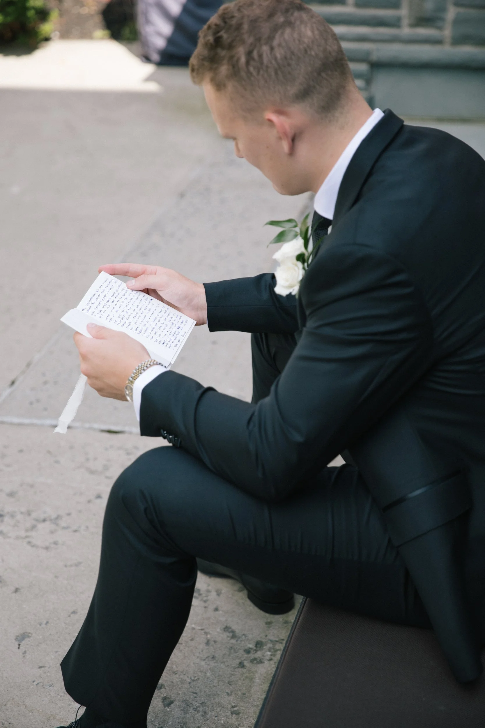 A man in a black suit with a white shirt and a boutonnière is sitting on a bench and reading a letter. He is wearing a wedding ring and watch, and is outdoors near some stairs and greenery.