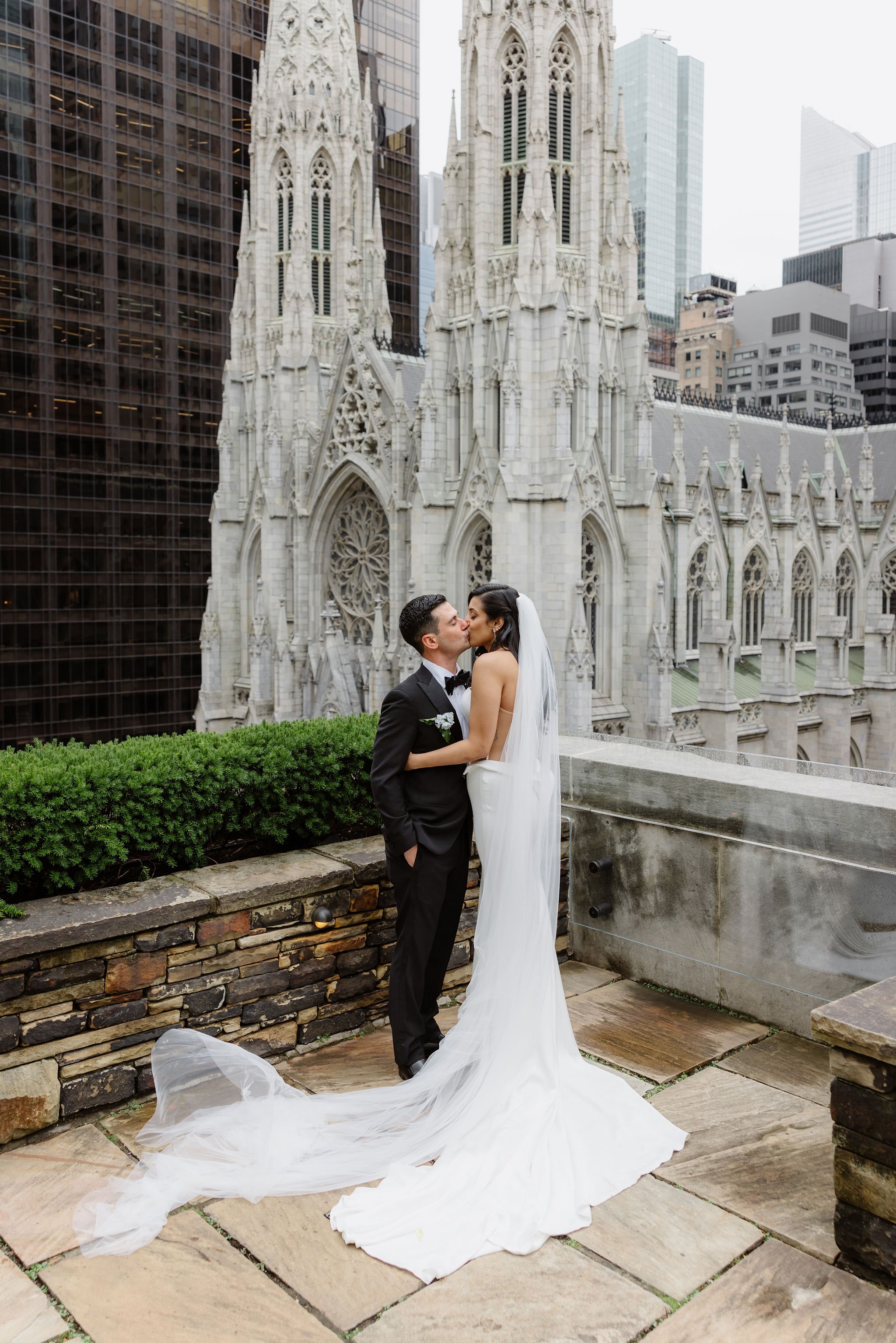 A newlywed couple sharing a kiss in front of a Gothic-style church with a cityscape of tall modern buildings behind them.