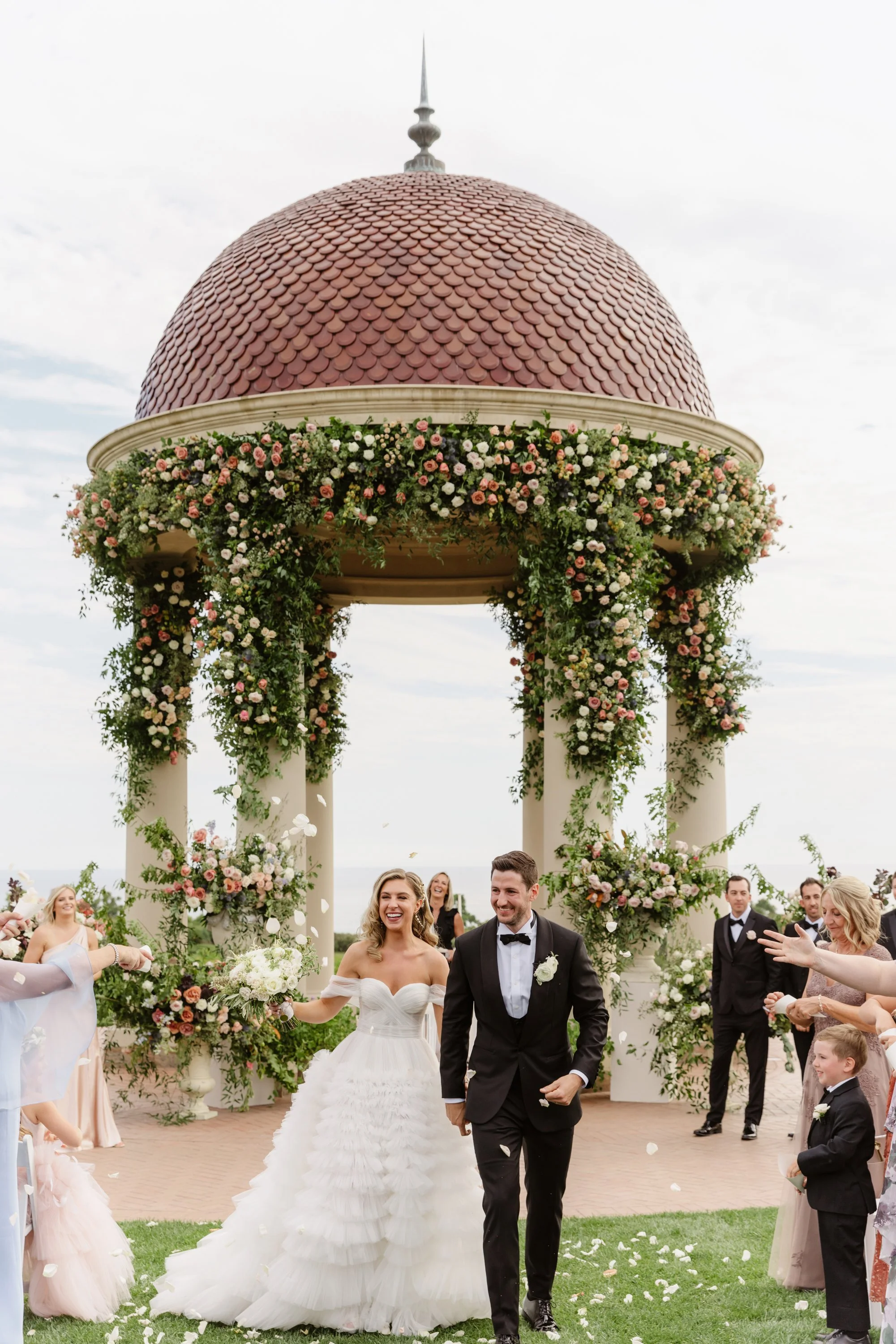 Wedding couple walking under a floral arch during ceremony with wedding guests around.