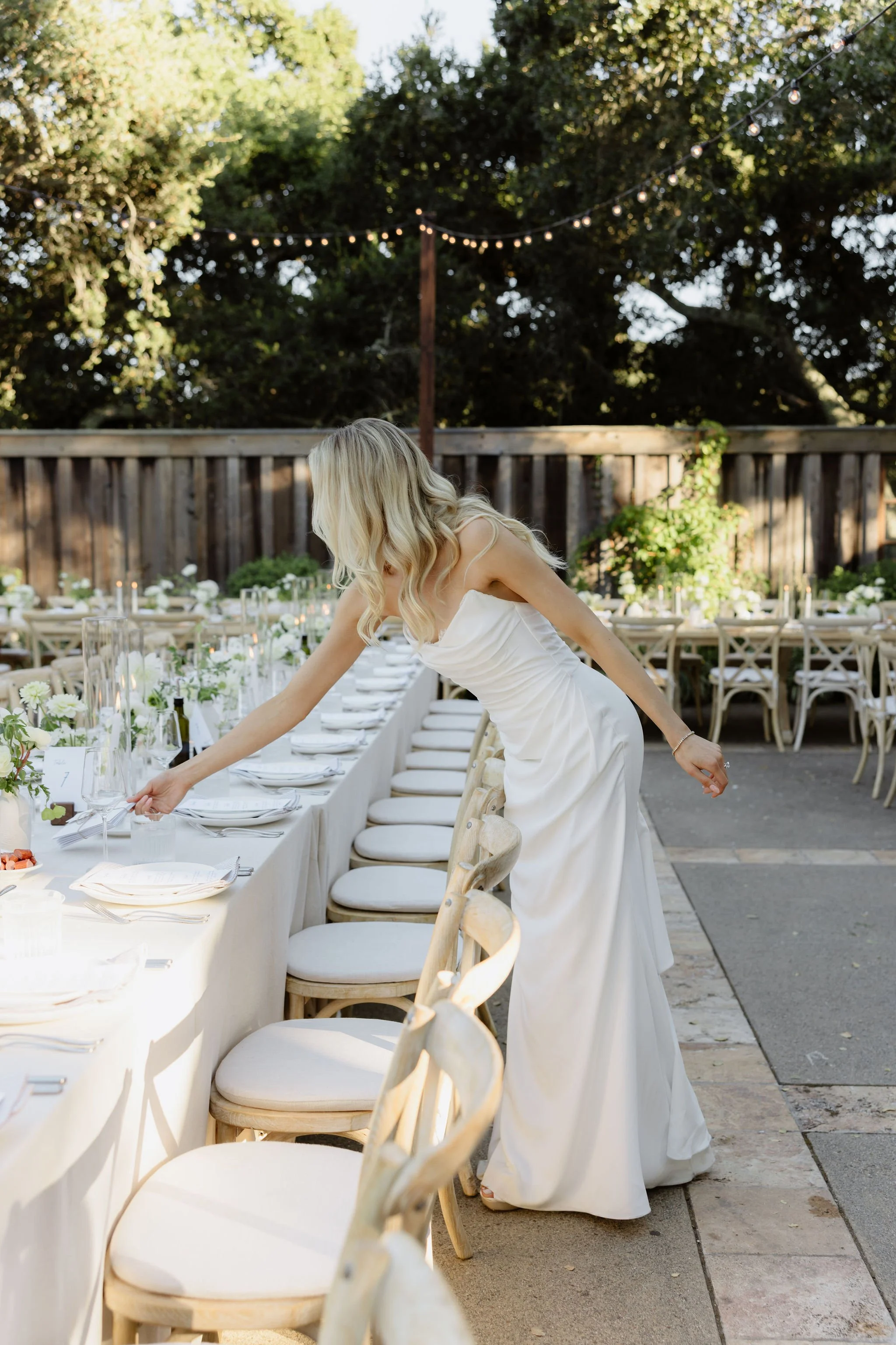 A woman in a white dress arranging a long banquet table with white tablecloth and floral centerpiece outdoors, with string lights overhead and a wooden fence in the background.