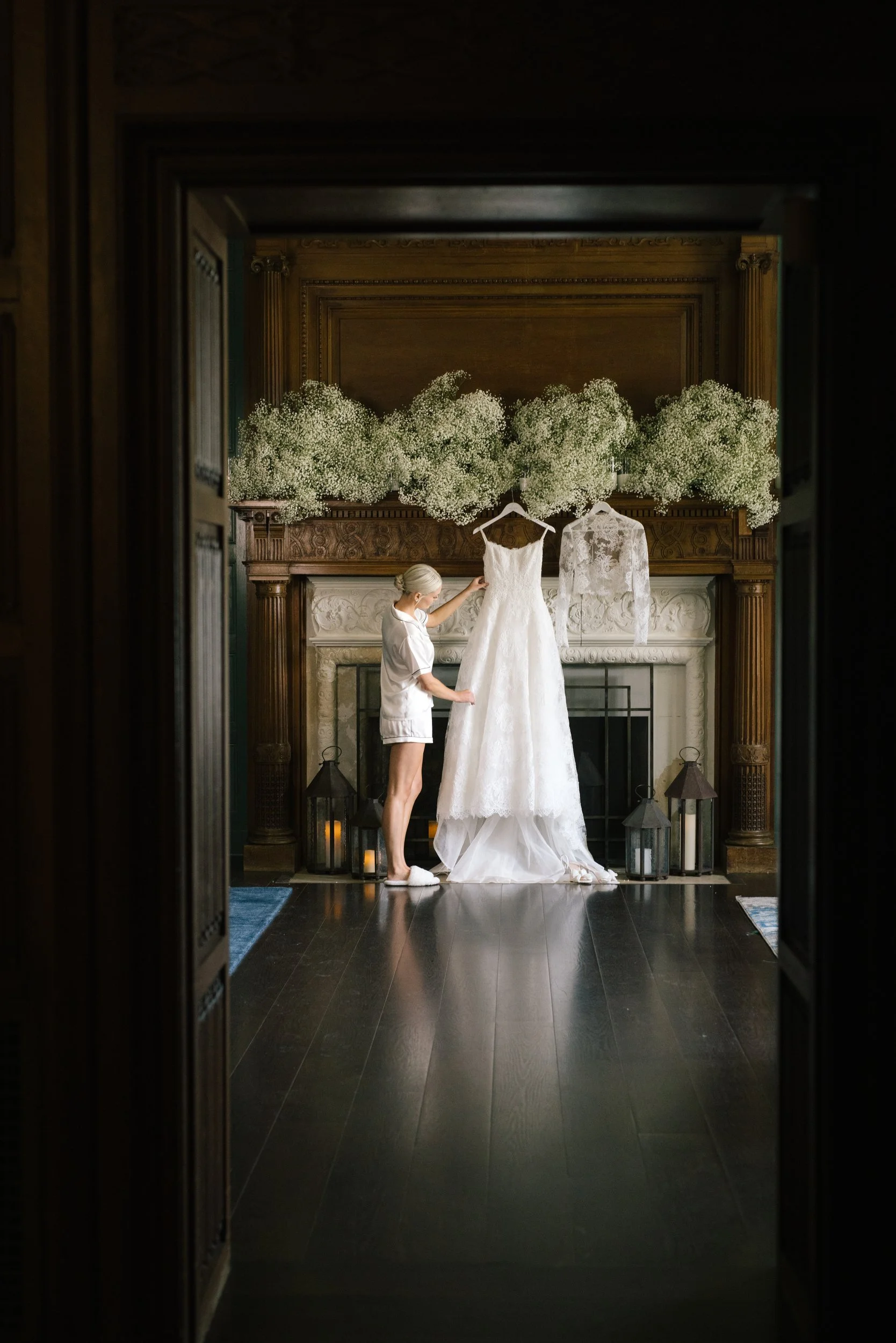 A woman with platinum blonde hair in a bun adjusts a white wedding dress hanging on a wooden fire surround, with large bouquets of white baby's breath flowers on top, in a dimly lit room with candles in lanterns on the floor.