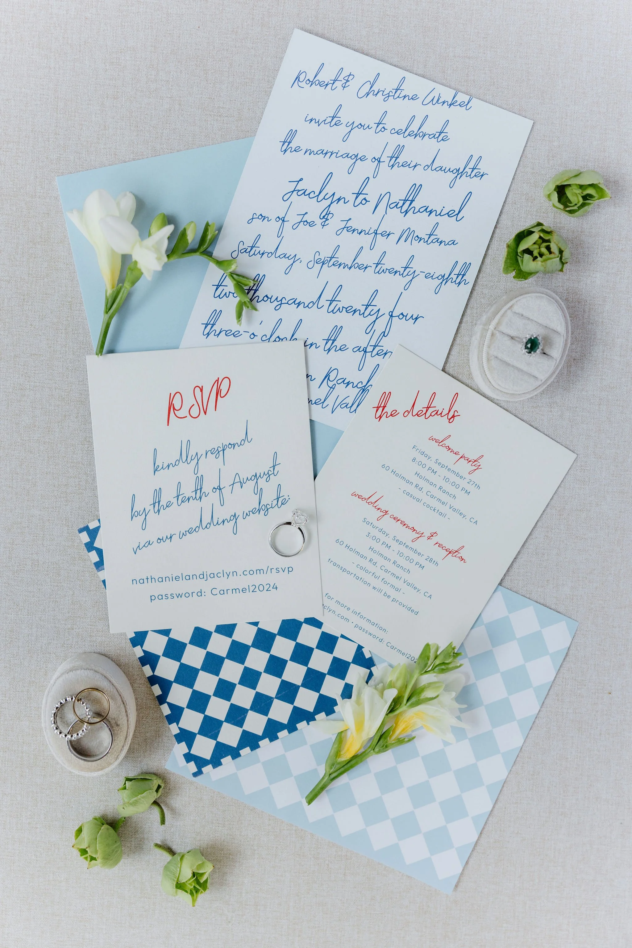 Wedding invitation, RSVP card, and details card with blue and red text, flowers, and rings on a light fabric background.