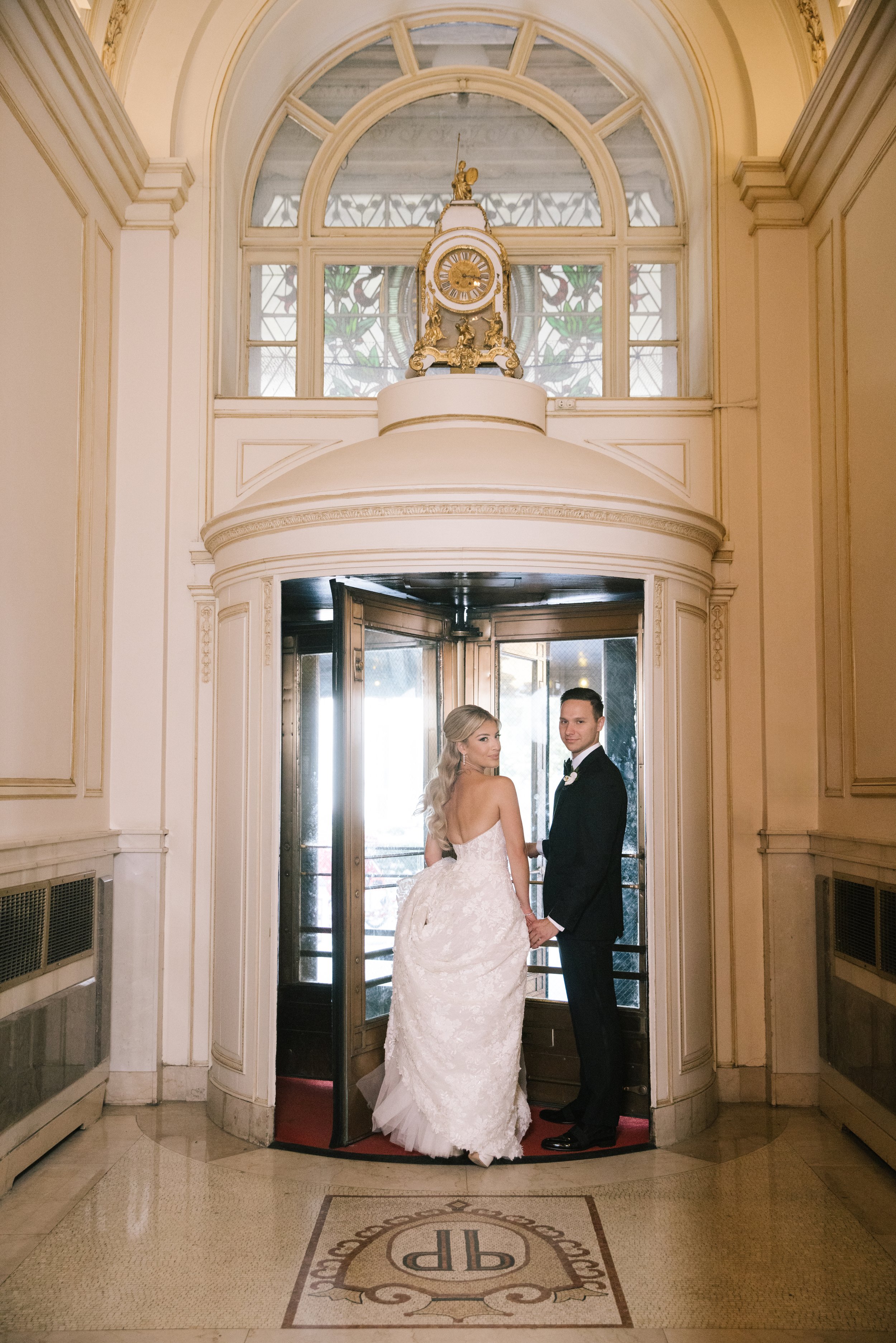 A bride and groom standing hand in hand inside a building with a grand entrance, ornate clock, stained glass window, and elegant architecture.