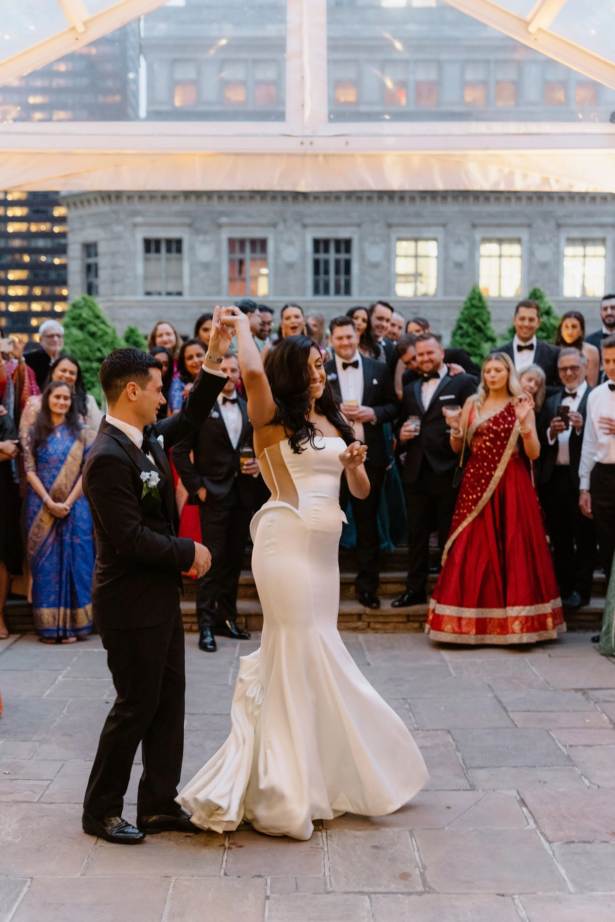 A wedding reception with the bride and groom dancing in front of a group of guests. The bride wears a white strapless wedding gown, and the groom is in a black tuxedo. Guests are dressed in formal attire including tuxedos and traditional Indian saree