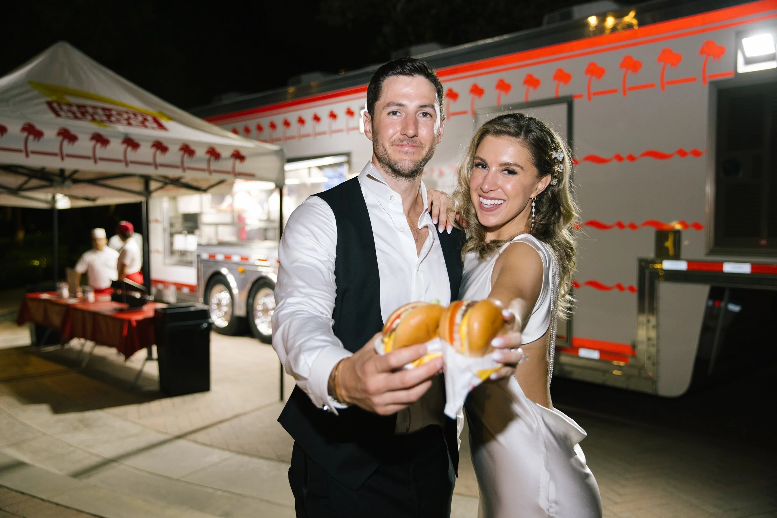 A man and woman dressed in formal attire at night, smiling and holding hot dogs, standing in front of a food truck with palm tree graphics and an outdoor dining area.