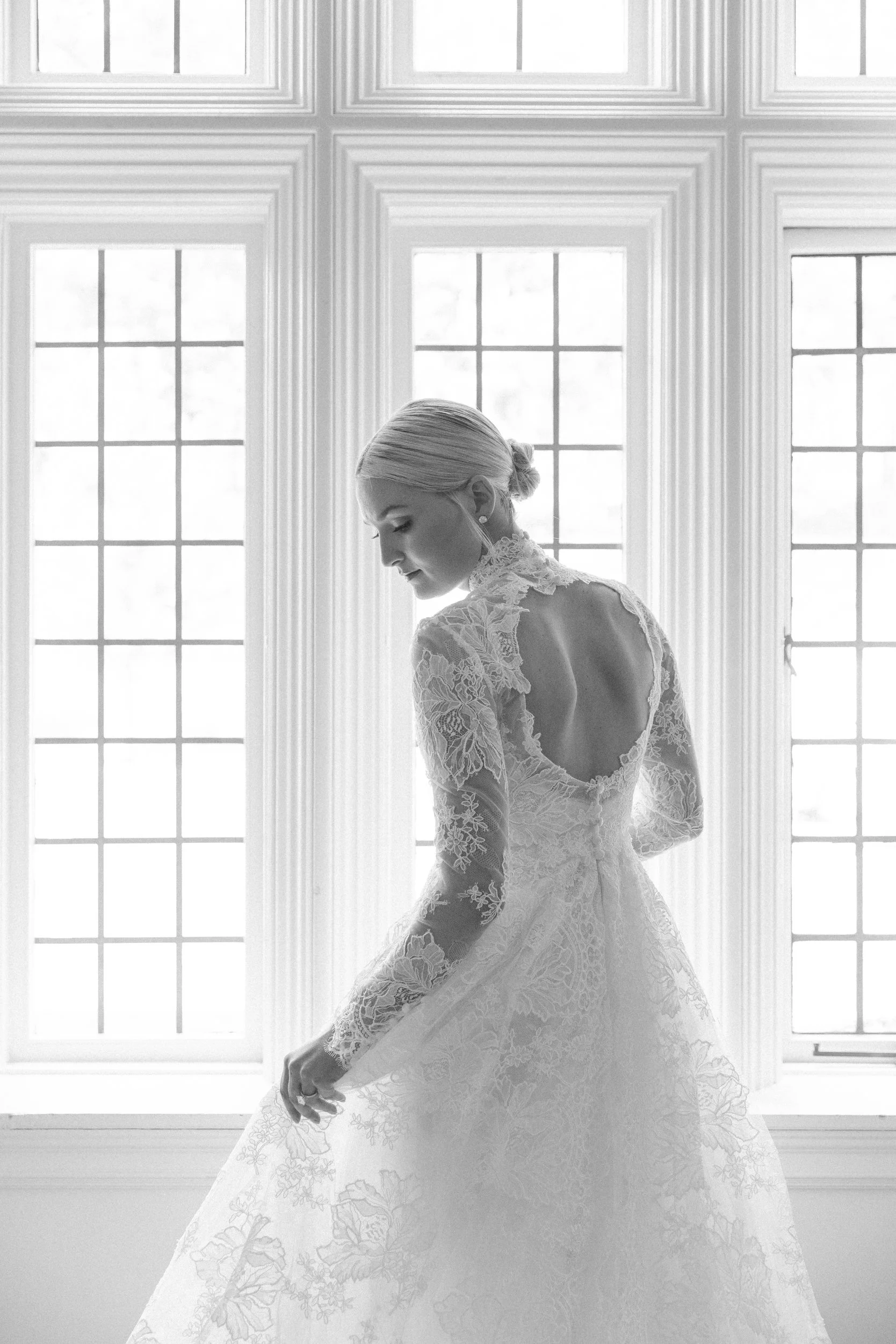 Black and white photo of a woman in a lace wedding dress looking down near large windows.