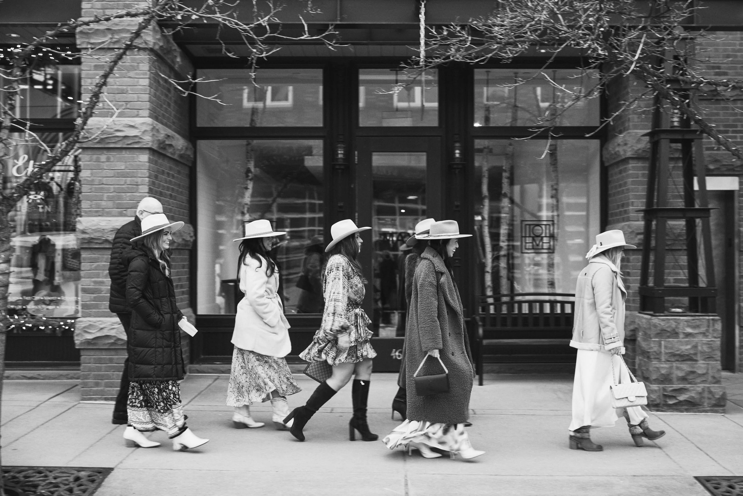 A group of women wearing fashionable clothing and wide-brimmed hats walking in a line on a city sidewalk in front of a brick building with large windows.