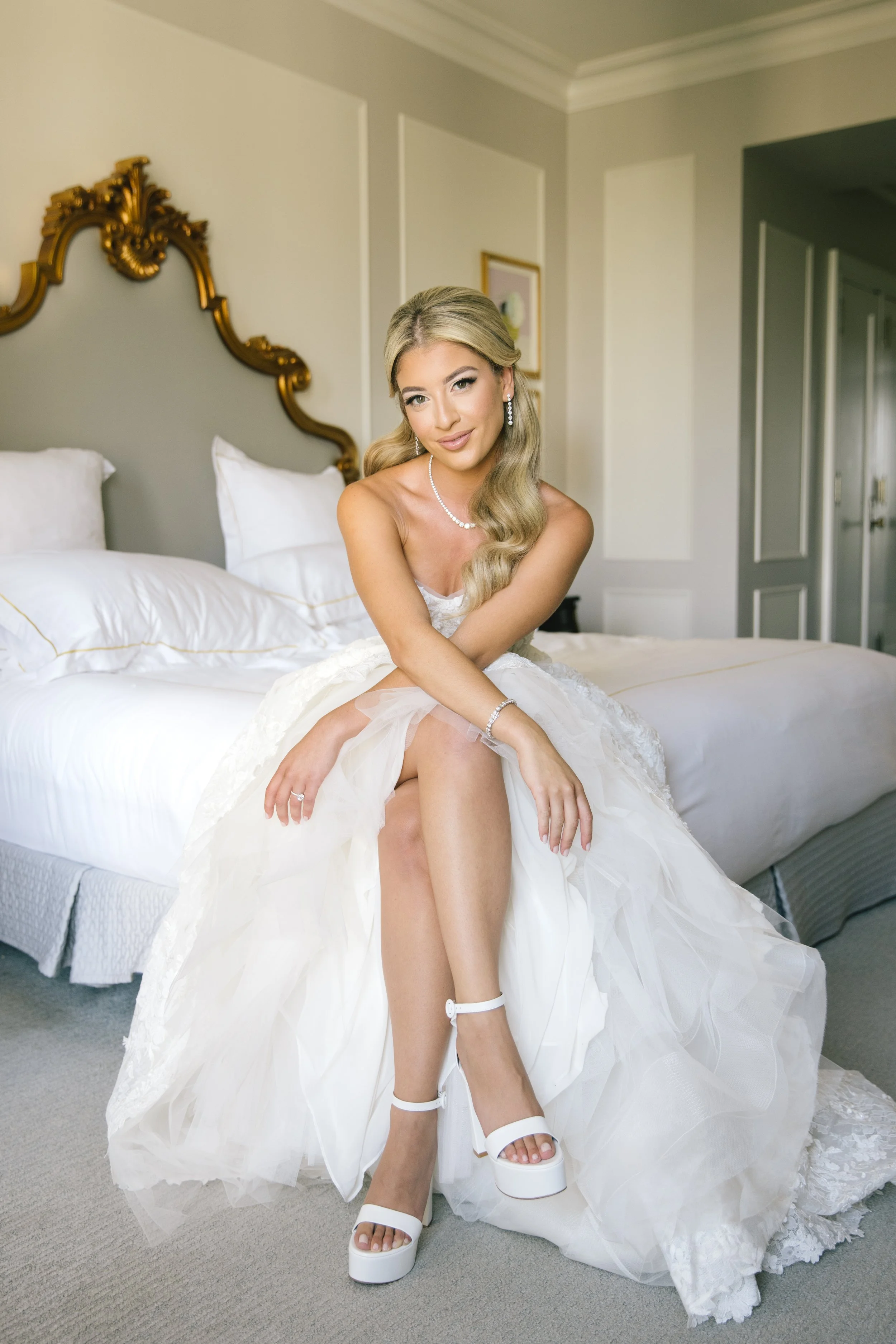A young woman in a wedding dress sitting on a bed with her feet partially visible, wearing white platform sandals, in a hotel room with ornate headboard and framed pictures on the wall.