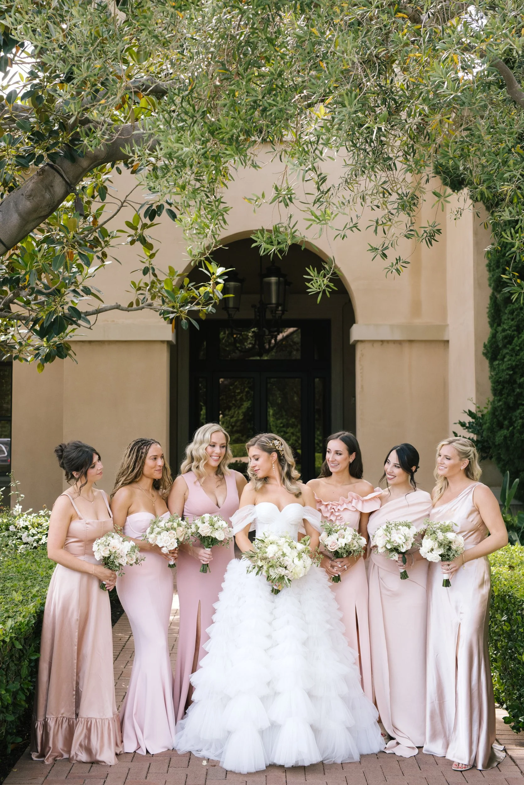 A bride and her bridesmaids pose outside a building with an arched entrance and hanging lanterns. The women wear matching blush pink dresses and hold floral bouquets. Greenery surrounds them, and a large tree with overhanging branches is above.