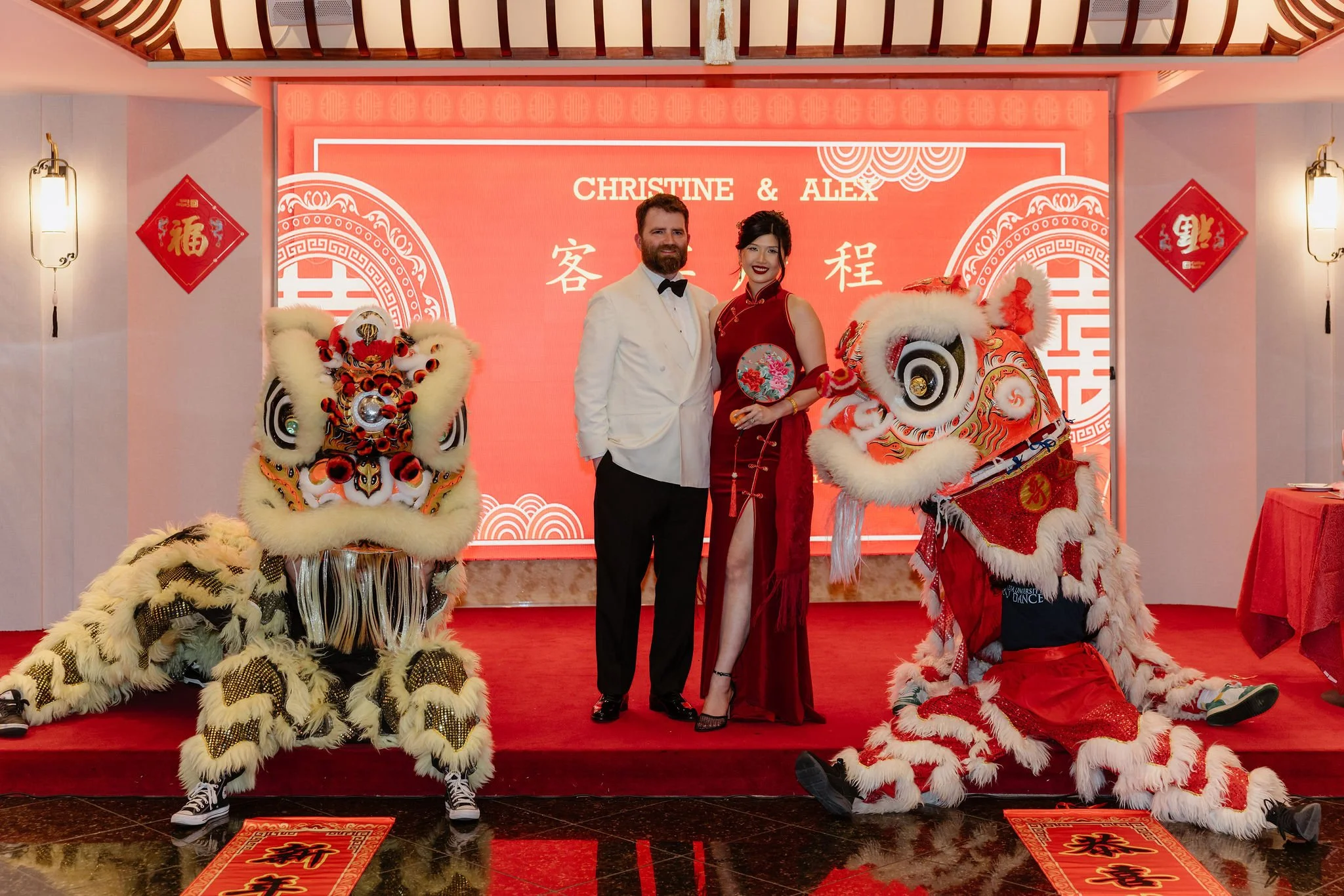 A couple stands on a red carpet at a Chinese celebration, flanked by two lion dance costumes. The background features a large red digital display with the names 'Christine & Alex' and Chinese characters. Traditional red and gold decorations adorn the