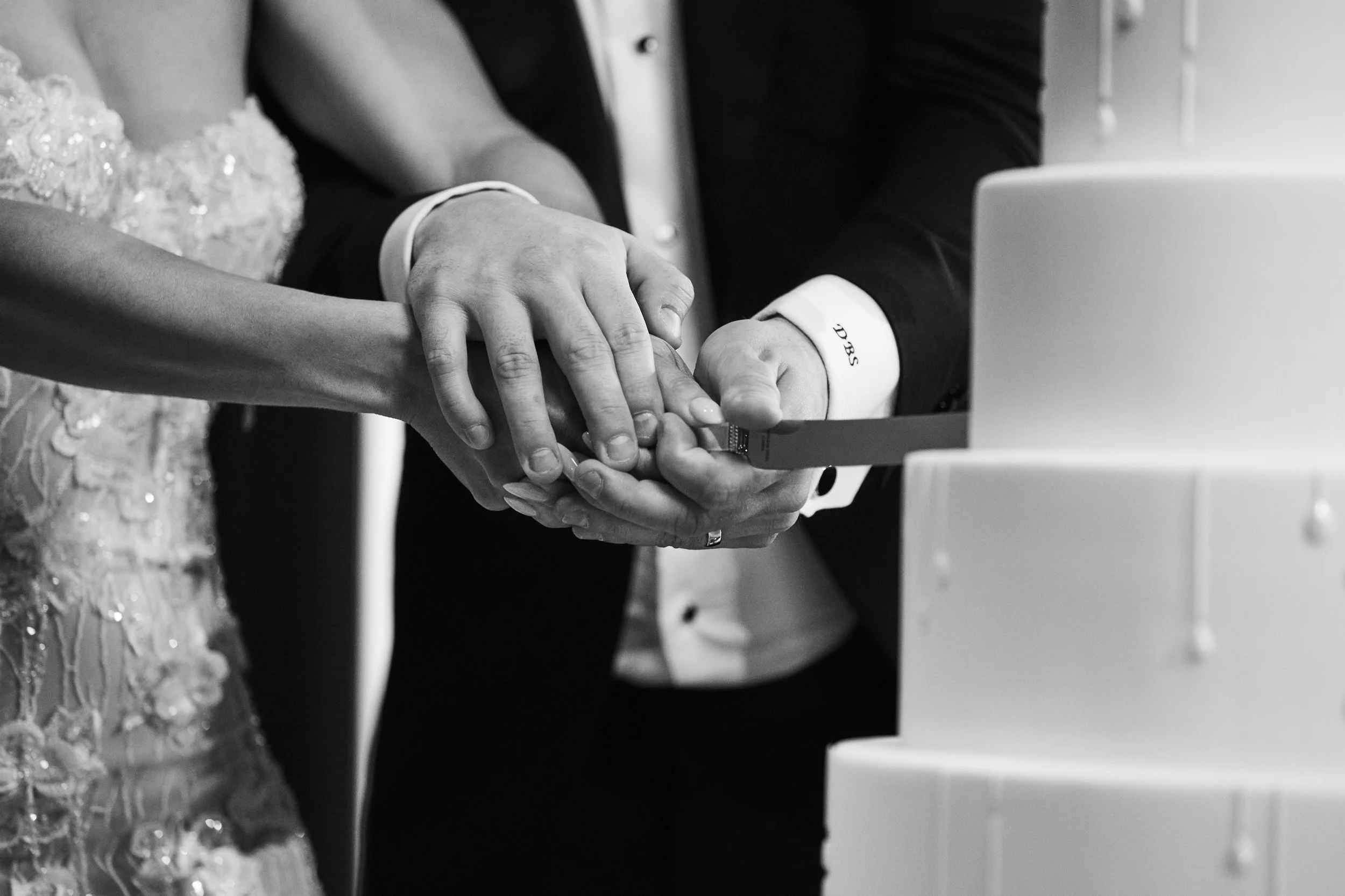 Black and white image of a couple holding hands during a wedding or formal event, with part of a wedding dress visible on the left and a laptop on the right.