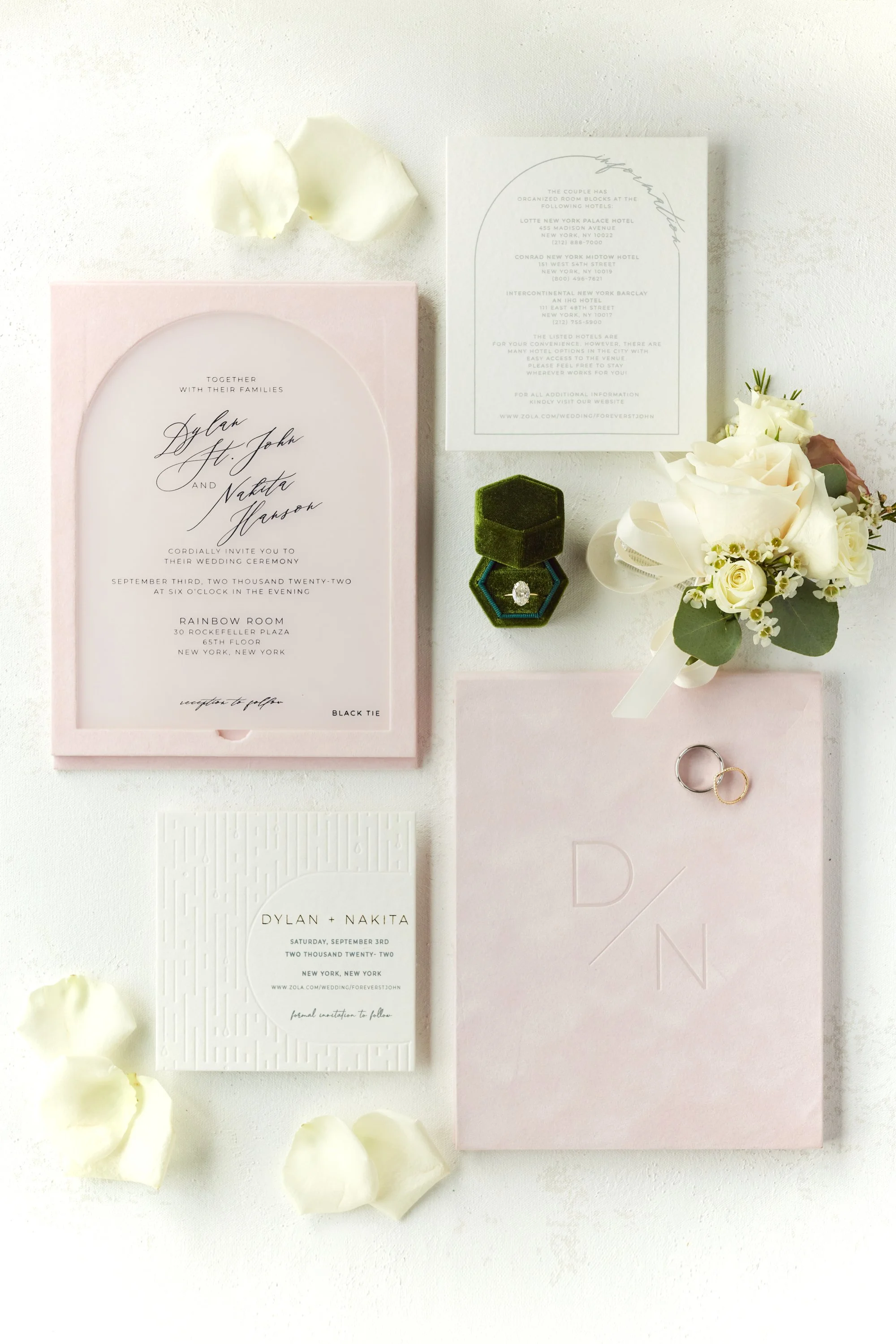 Wedding invitation suite including an invitation card, RSVP card, floral boutonniere, and wedding rings, with scattered white flower petals on a white surface.