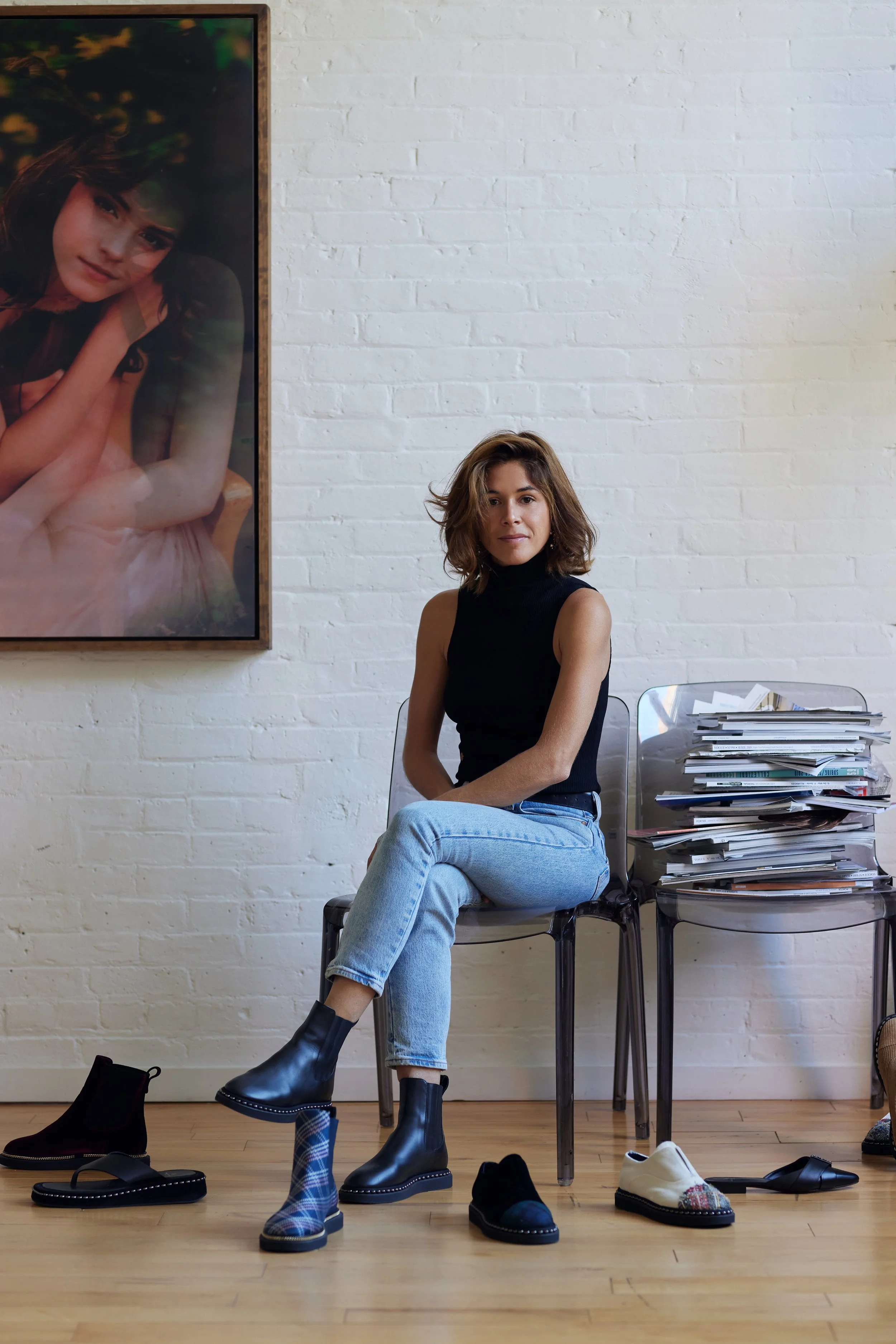 A woman with shoulder-length brown hair wearing a black sleeveless turtleneck and light blue jeans is sitting on a clear chair in a room with a white brick wall. She has her arms crossed and is looking at the camera. Behind her is a stack of magazine
