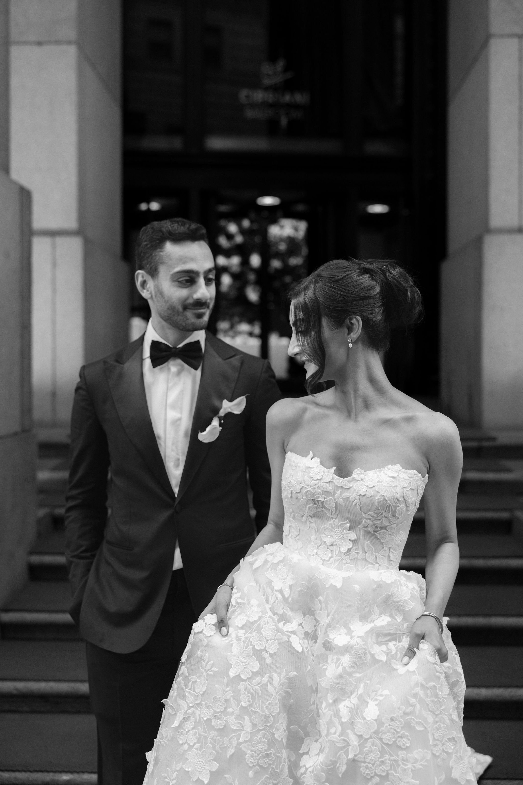 A black and white photo of a bride and groom standing close together outside a building, smiling at each other. The groom is wearing a tuxedo with a bow tie, and the bride is wearing a strapless wedding dress with lace detailing, holding the skirt wi