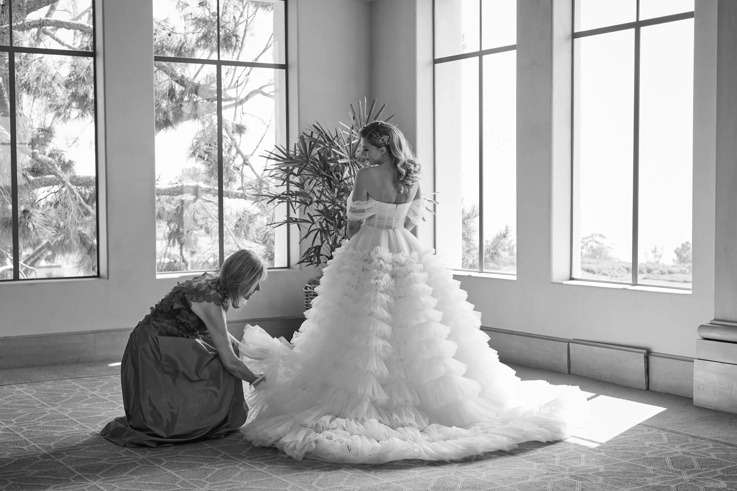 A bride in a strapless wedding dress stands in front of large windows, with one woman kneeling and helping her with the dress. The bride's dress has a layered, ruffled skirt, and she has wavy hair with a hairpiece. There is a large indoor plant behin