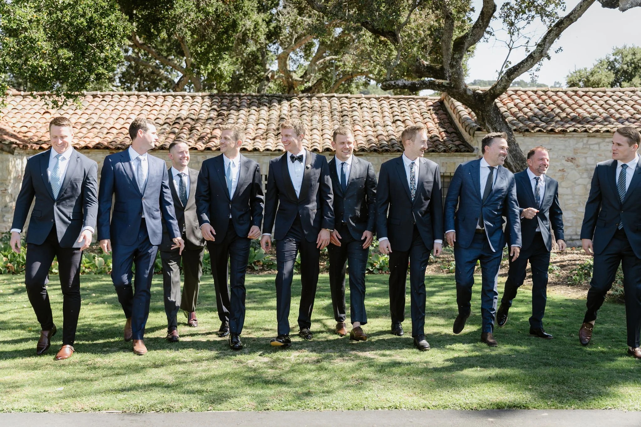 A group of men in suits walking outdoors on a sunny day, with a stone building and large trees in the background.