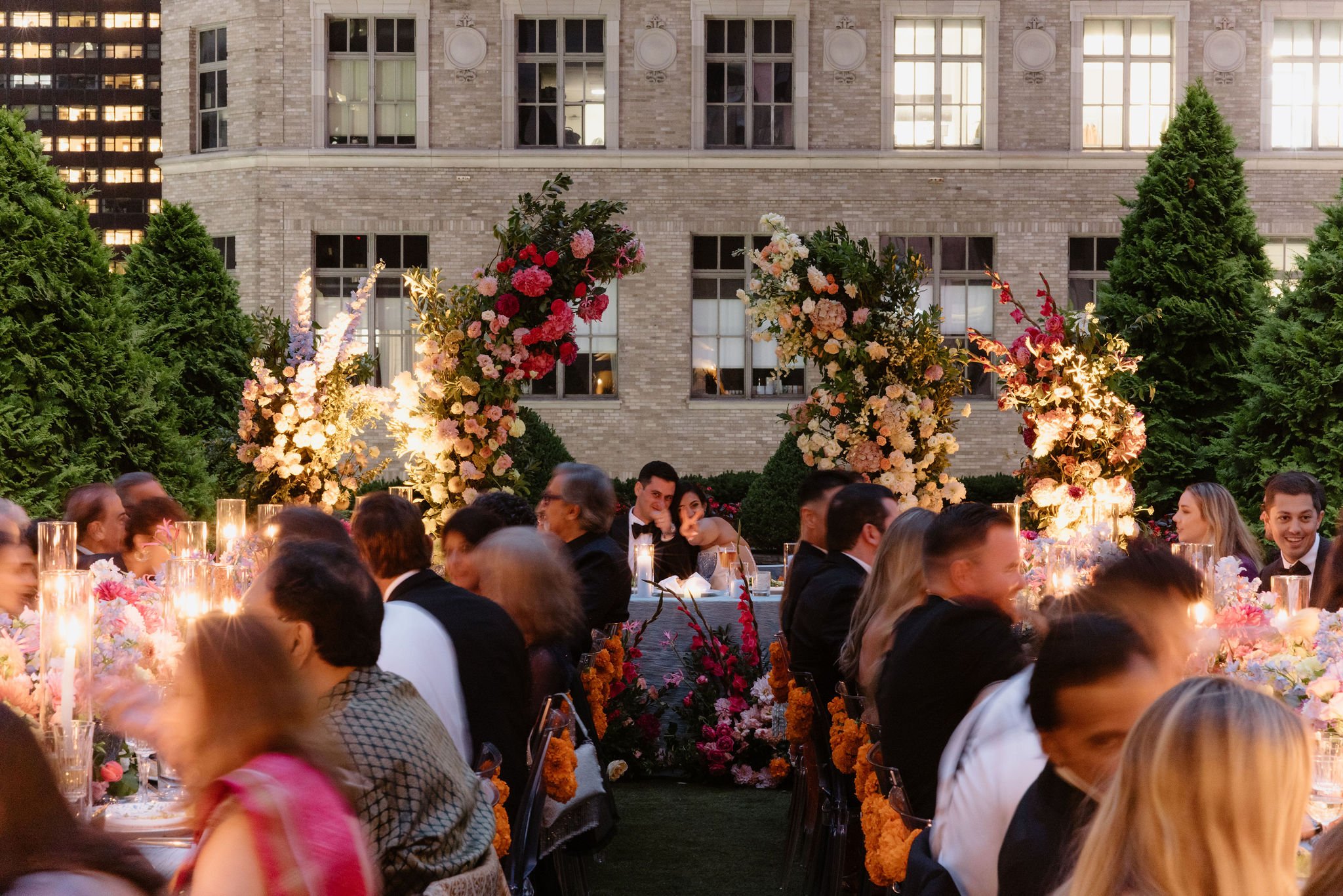 A nighttime outdoor gathering with people seated at tables, decorated with abundant floral arrangements and candles, in front of a brick building with lit windows.