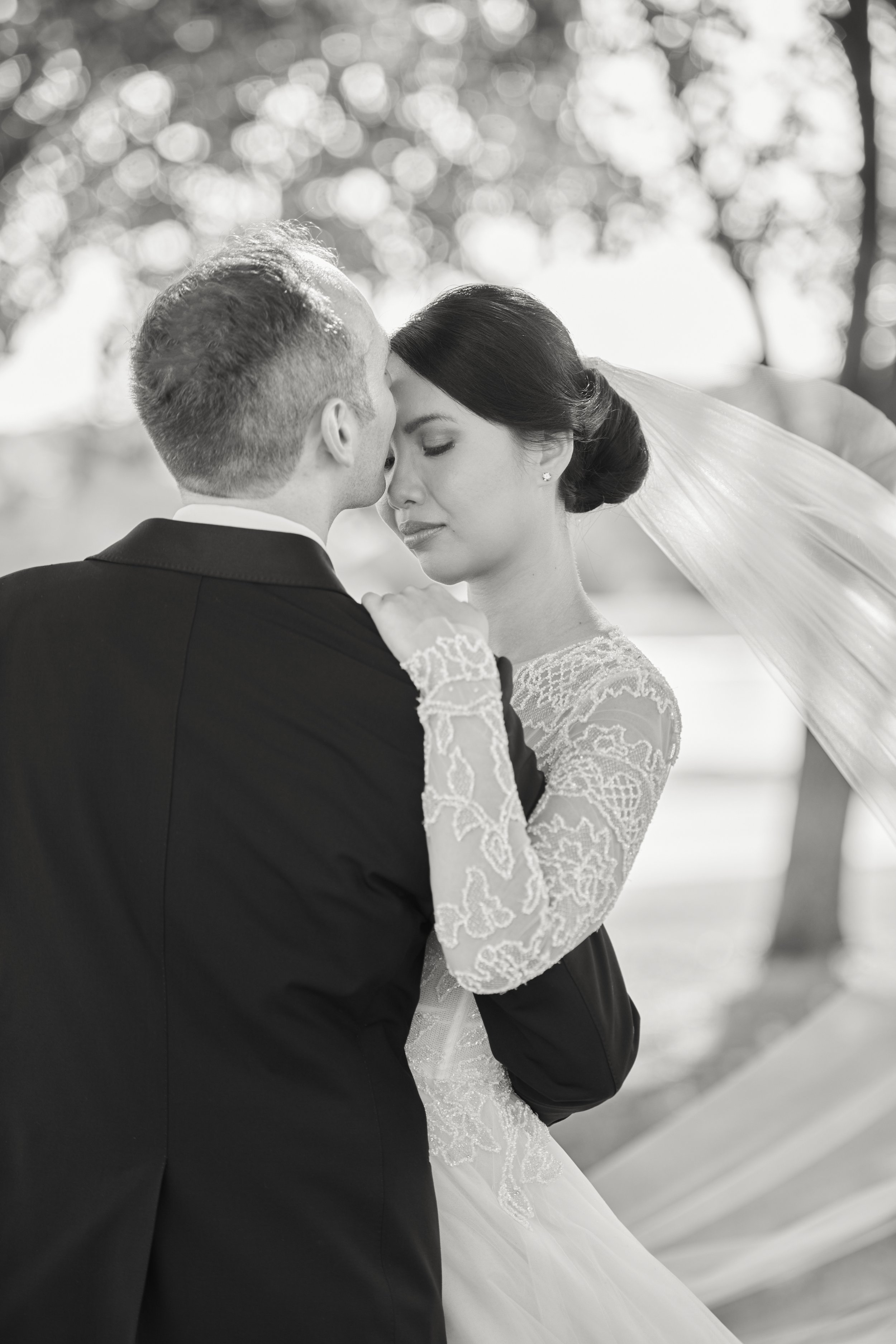 A black-and-white photo of a bride and groom embracing outdoors, with the groom's face close to the bride's forehead and her eyes closed. The couple is dressed in wedding attire, with the bride wearing a lace dress and veil, and the groom in a suit. The background is blurred with bokeh from sunlight filtering through trees.