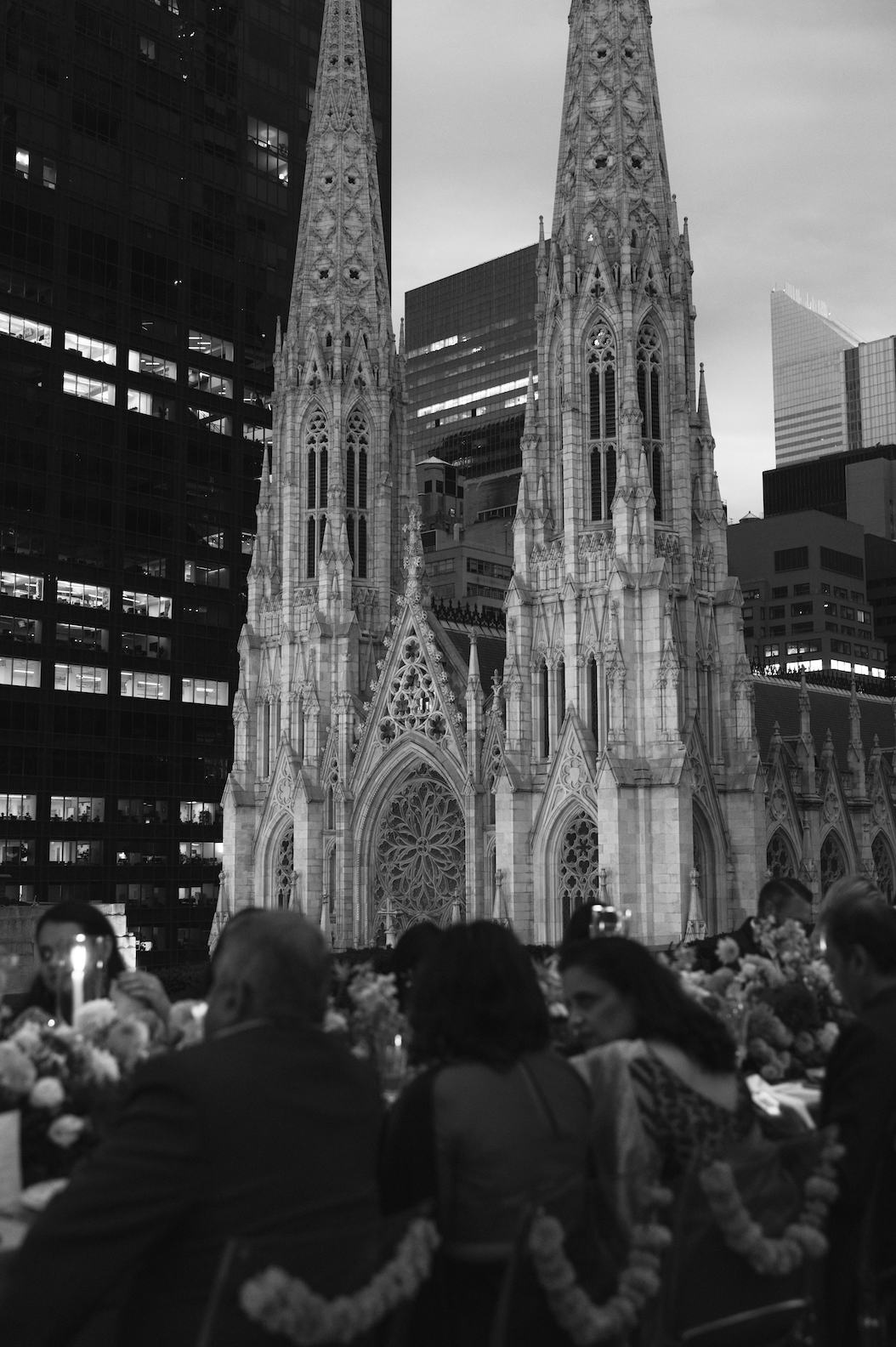 Black-and-white photo of a large cathedral with skyscrapers in the background, people sitting at tables, and floral decorations in the foreground.