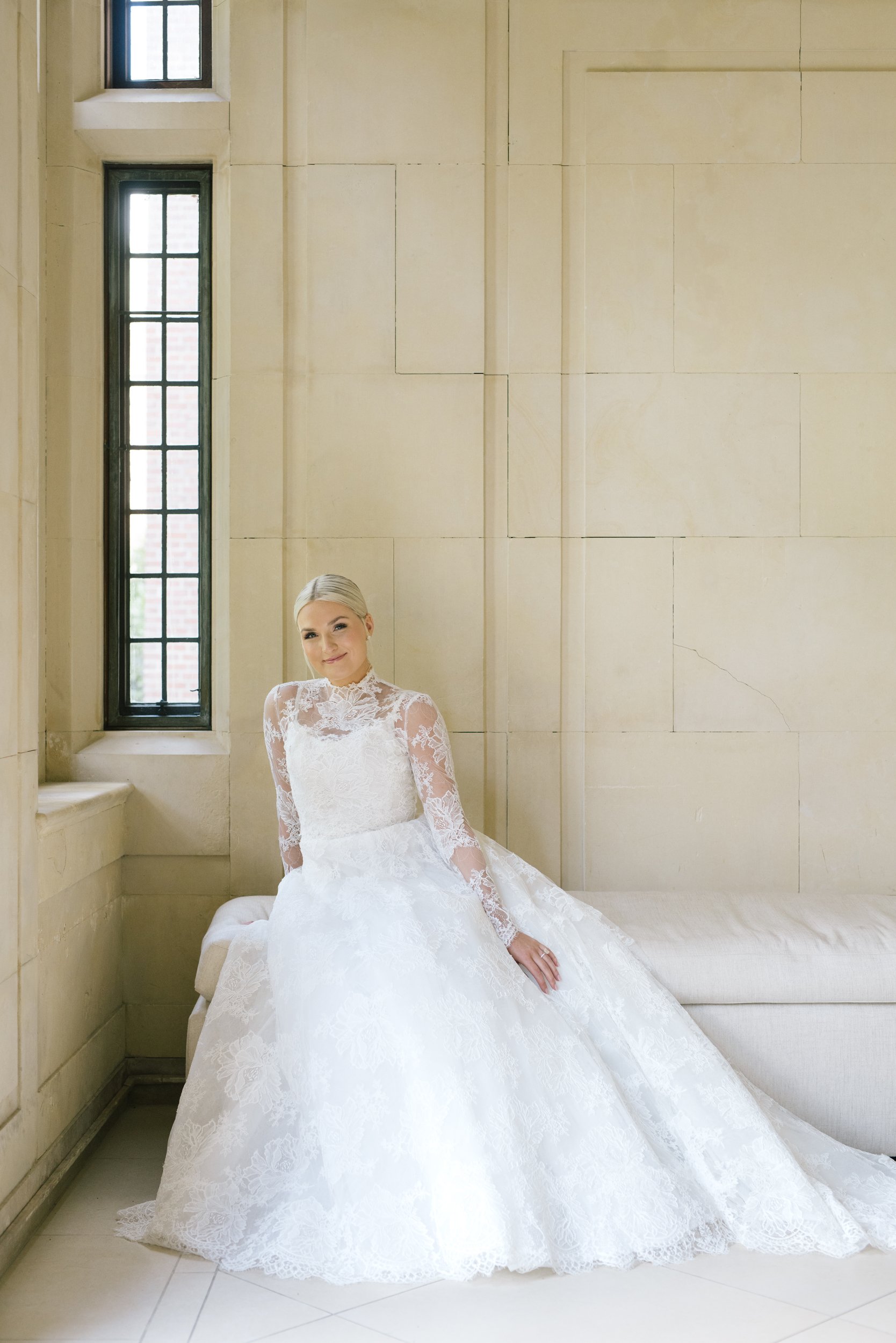 A bride sitting on a beige cushioned bench in front of a large beige wall with a tall narrow window on the left, wearing a white lace wedding dress with long sleeves and a full skirt.
